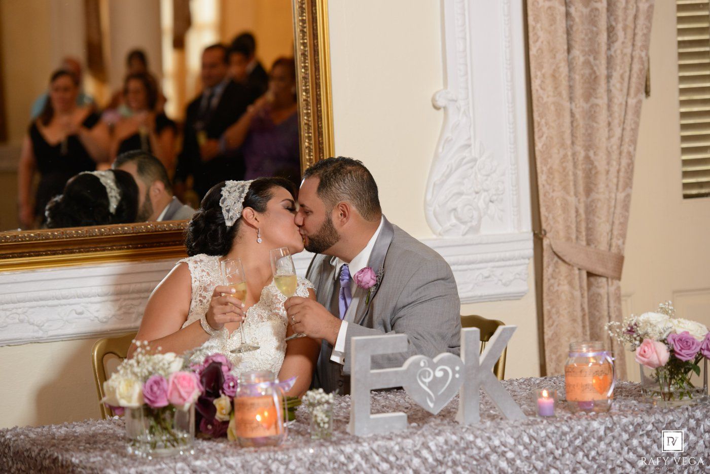 Newlyweds sharing a kiss at their wedding reception sweetheart table decorated with floral arrangements and monogram letters.
