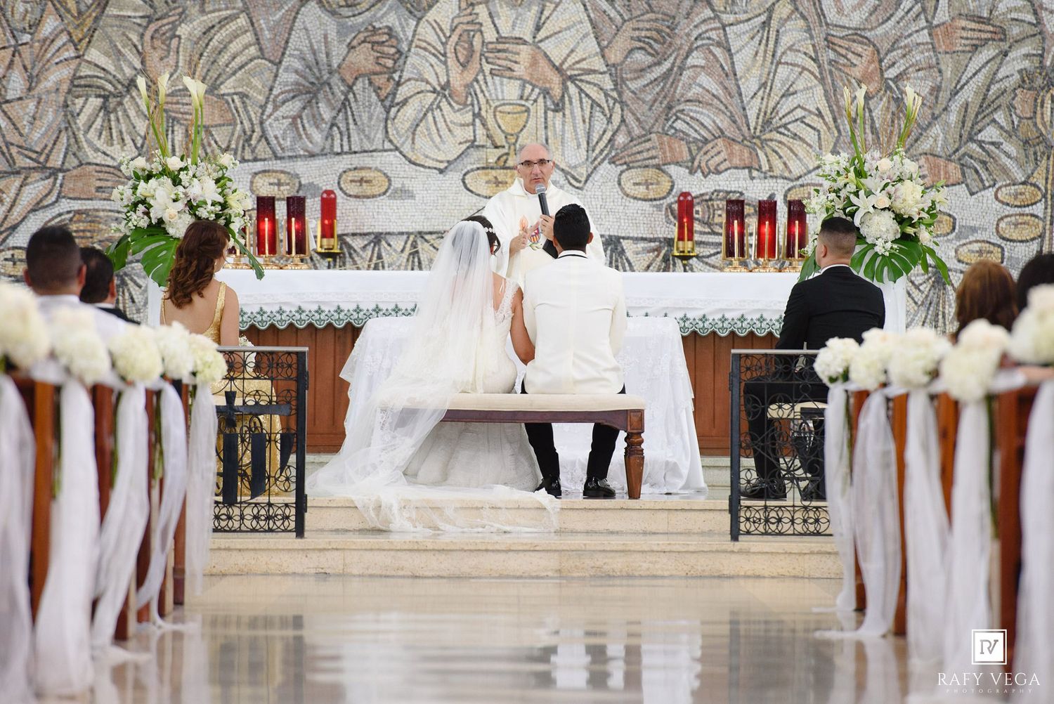 Elegant church wedding ceremony with bride and groom at altar, white floral arrangements, and officiant in white robes.