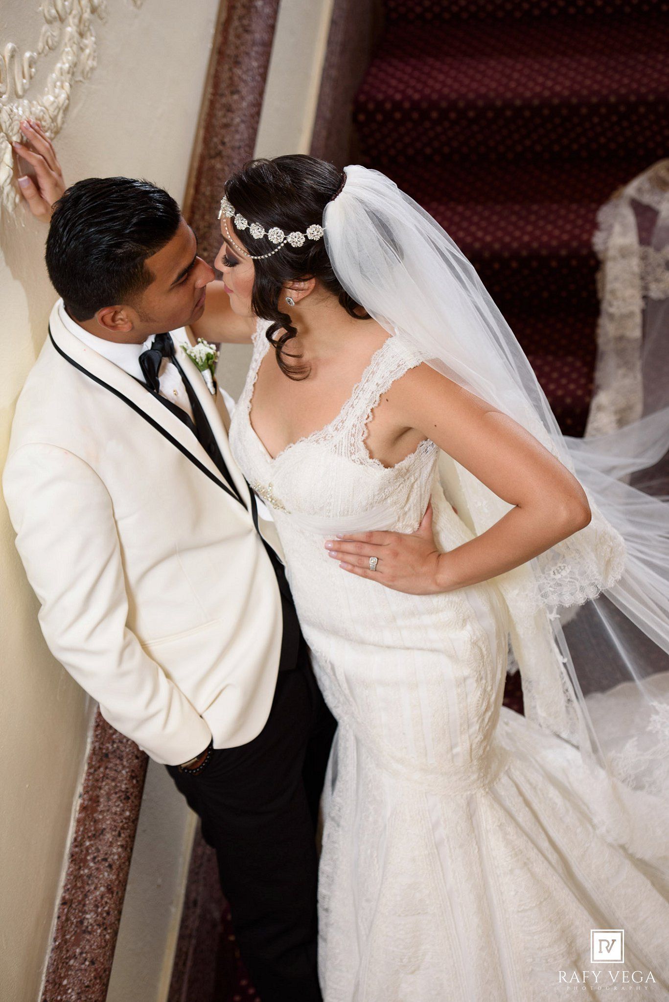 Bride and groom share a romantic kiss on a staircase, bride wearing a lace gown with veil and floral headpiece.