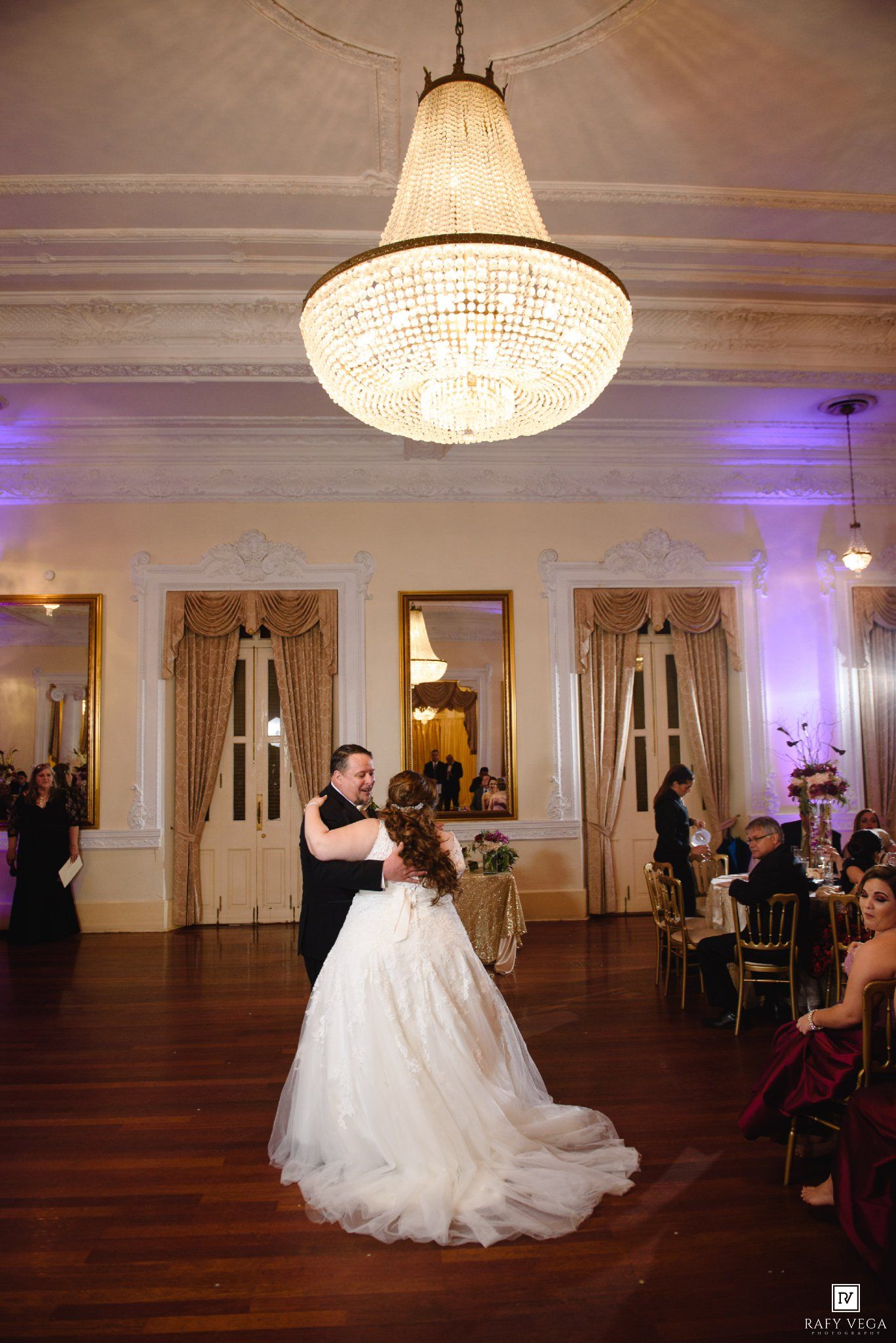 Bride and groom share first dance under crystal chandelier in elegant ballroom with ornate moldings and purple uplighting.
