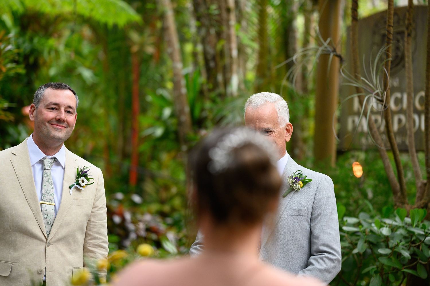 Two grooms smiling joyfully during an outdoor wedding ceremony surrounded by lush tropical greenery.