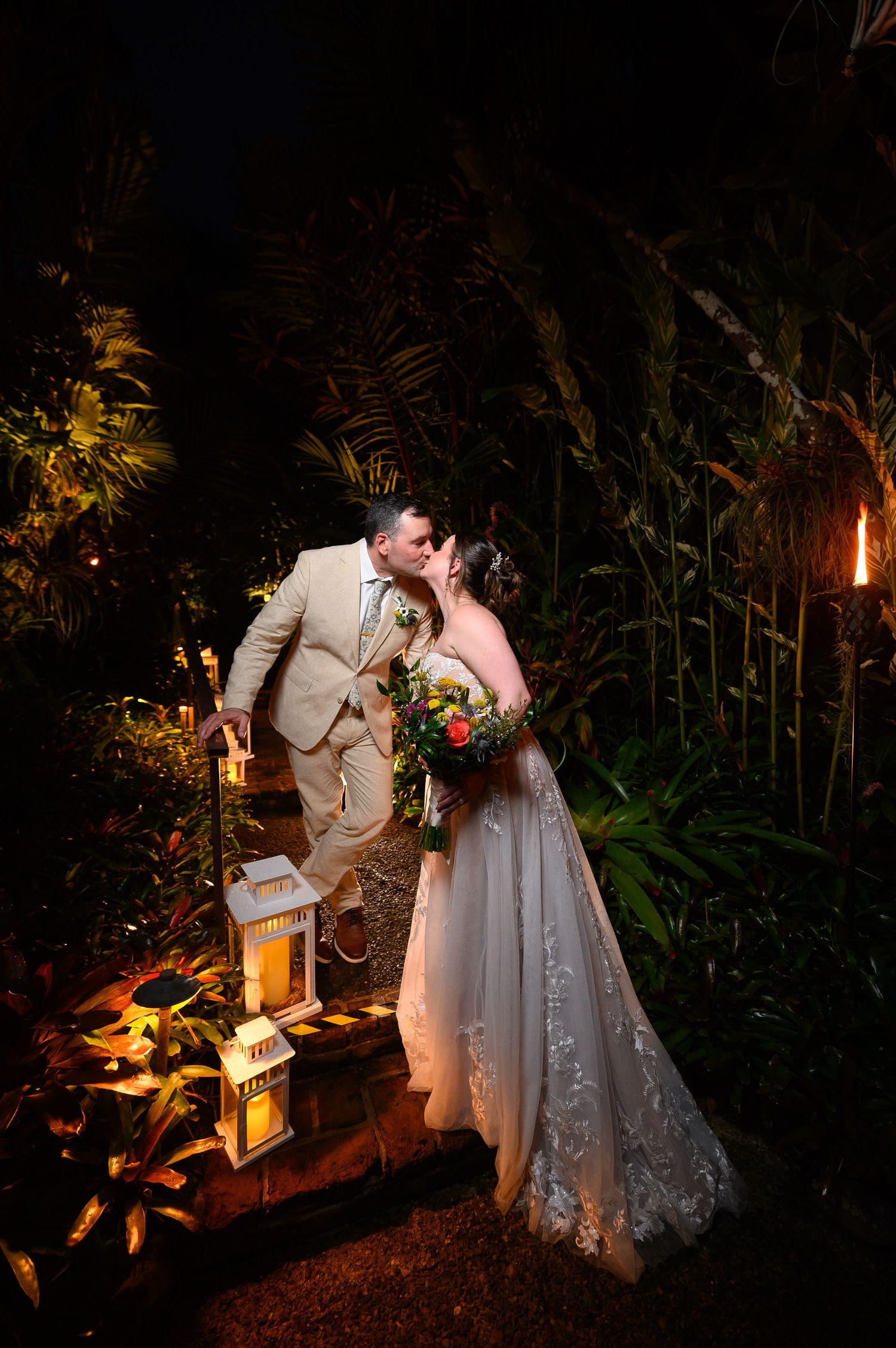 Bride and groom share a romantic kiss at night surrounded by tropical greenery and glowing lanterns.