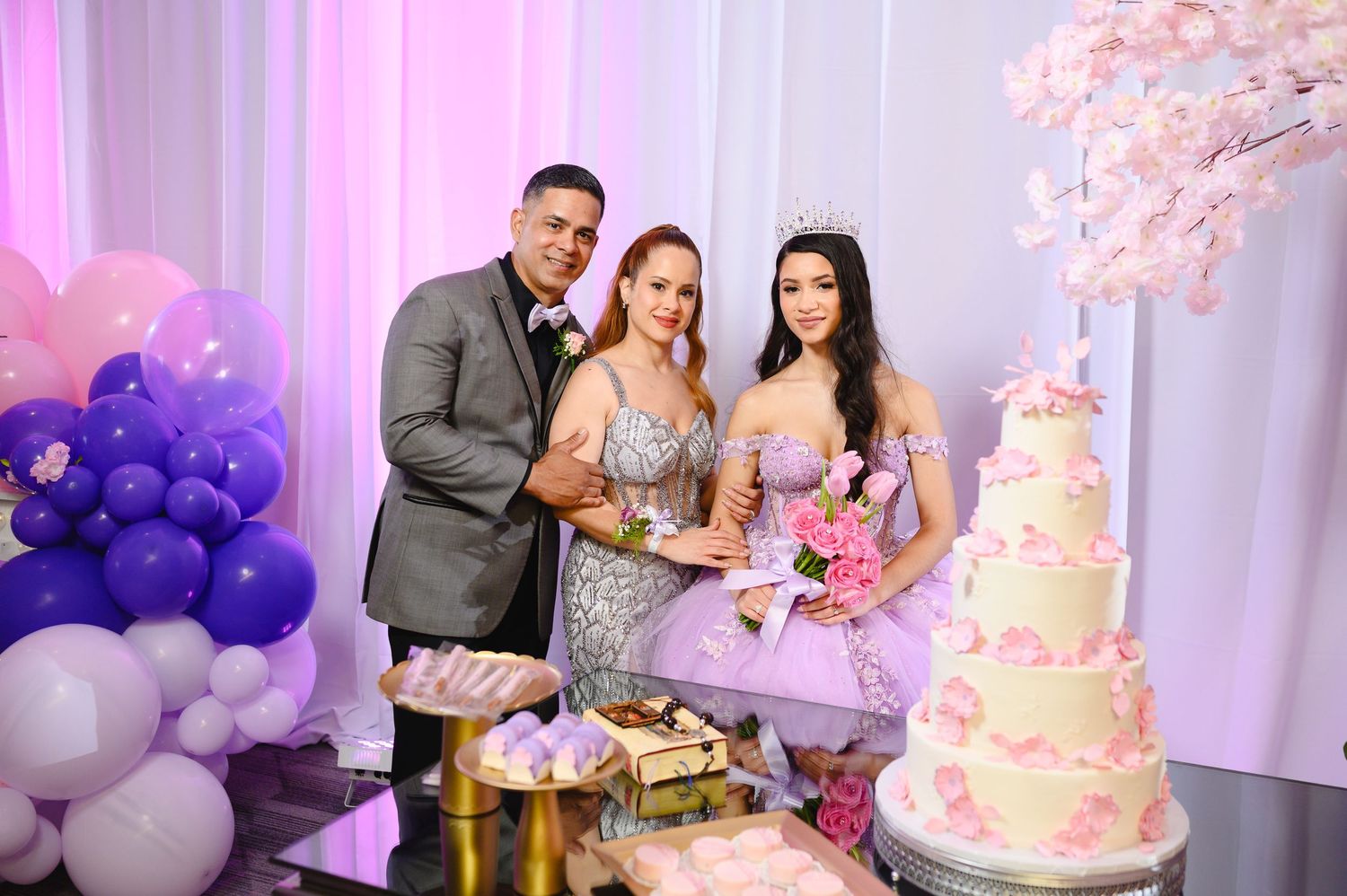 Family posing together at a quinceañera celebration with a pink floral cake, balloon decor, and the birthday girl in a tiara.
