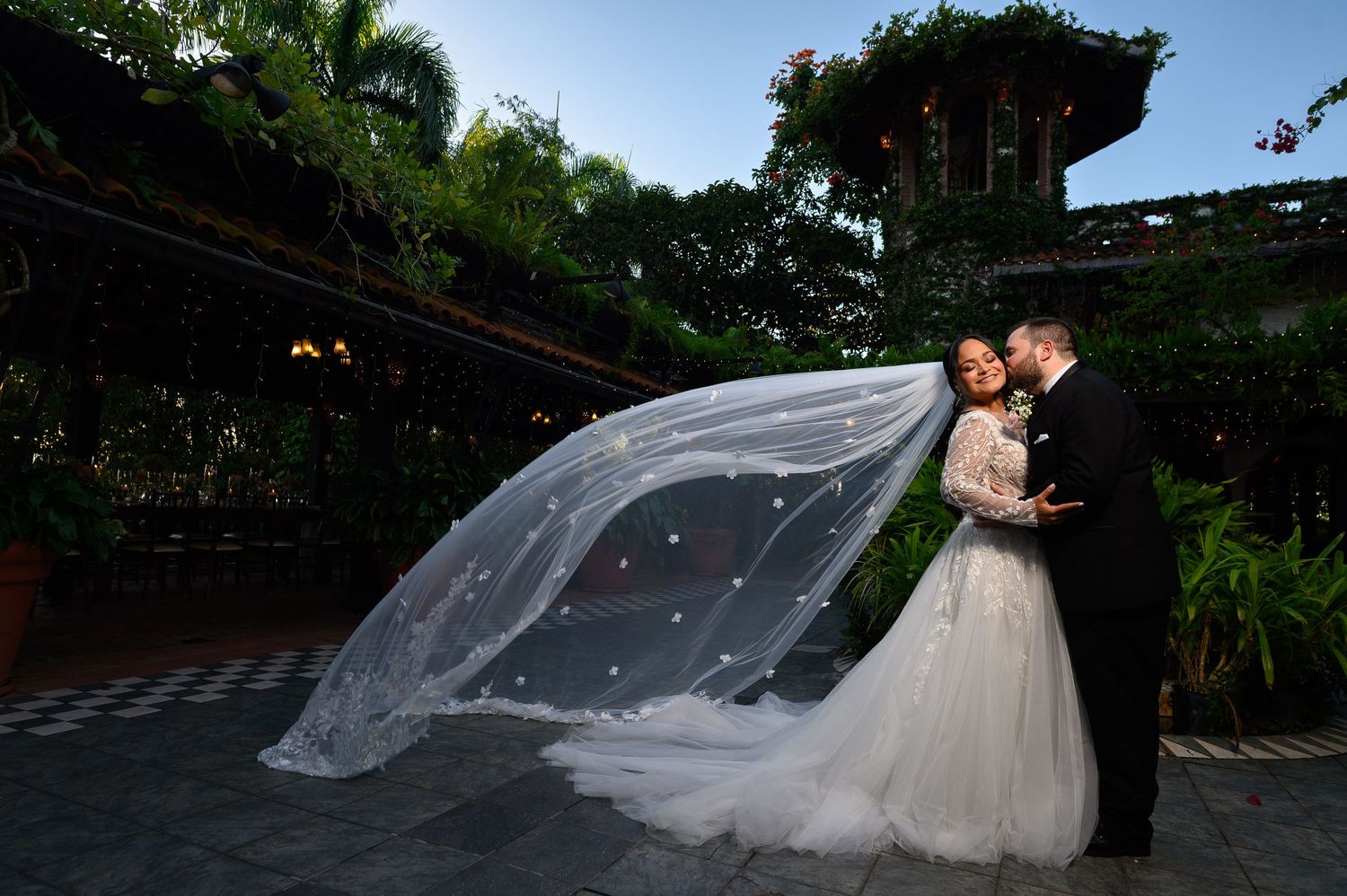 Bride and groom share romantic moment with flowing veil billowing in wind at lush garden wedding venue.