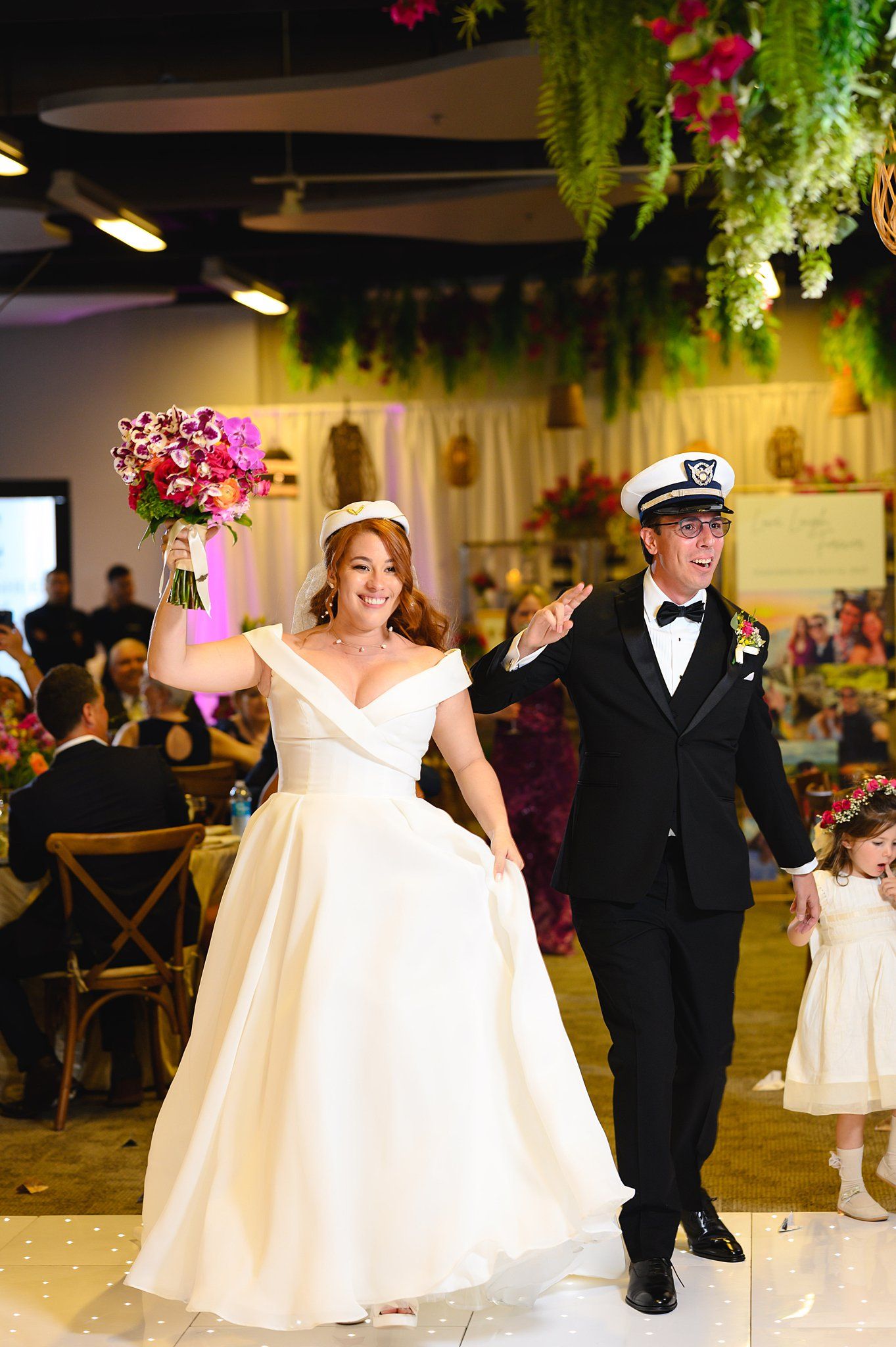 Bride in white gown raises bouquet as groom in sailor hat and tuxedo celebrate their wedding reception entrance.