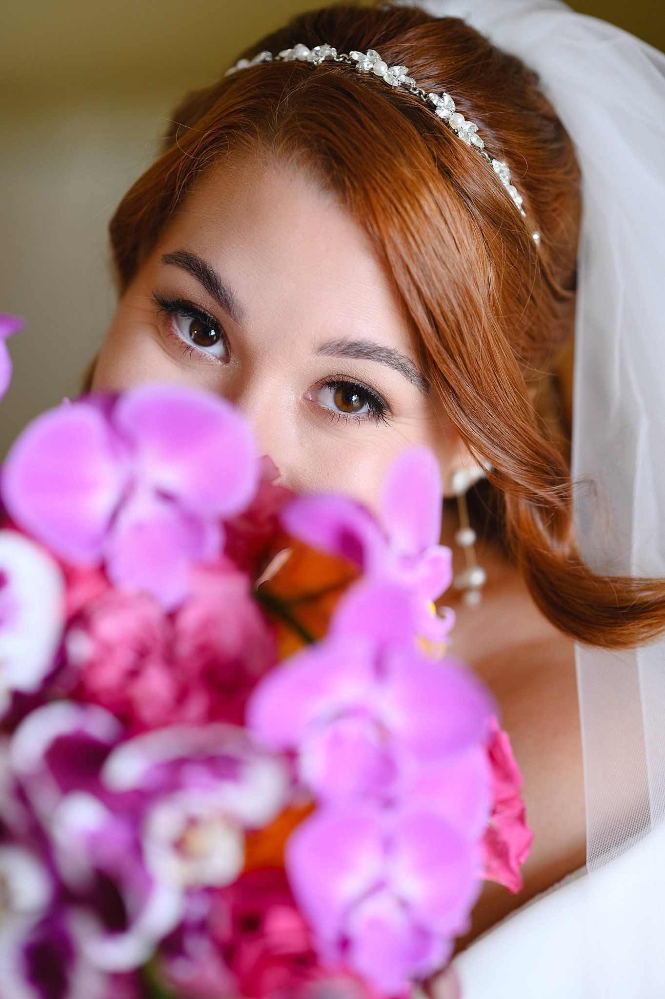 Bride holding vibrant pink orchid bouquet partially covering her face, wearing a veil and floral tiara.