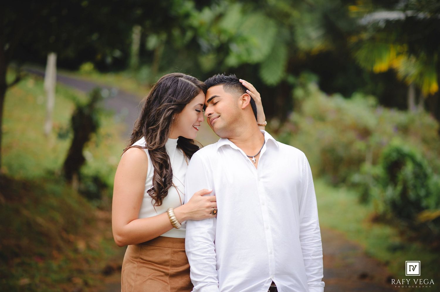 Couple sharing a romantic moment on a lush green path, woman in tan skirt embracing man in white shirt.