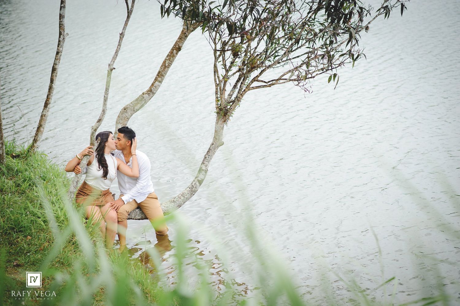 Couple sharing a romantic kiss by a serene lakeside, seated near trees on a grassy bank in a beautiful outdoor setting.