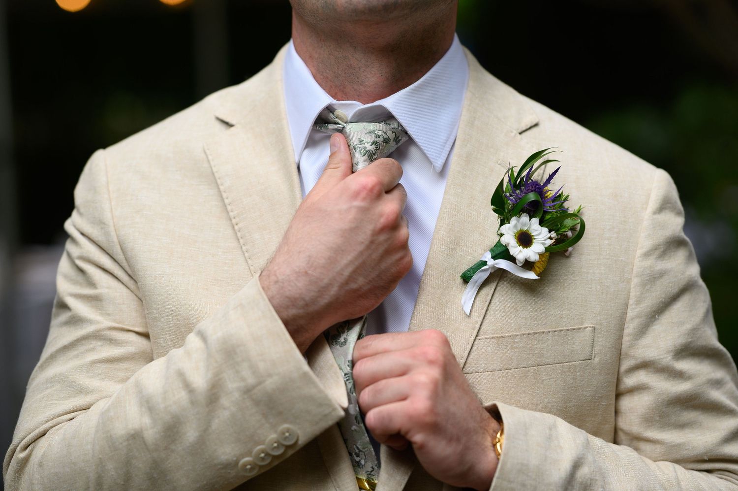 Man in beige suit adjusting tie with floral boutonniere featuring white daisy and purple flowers.