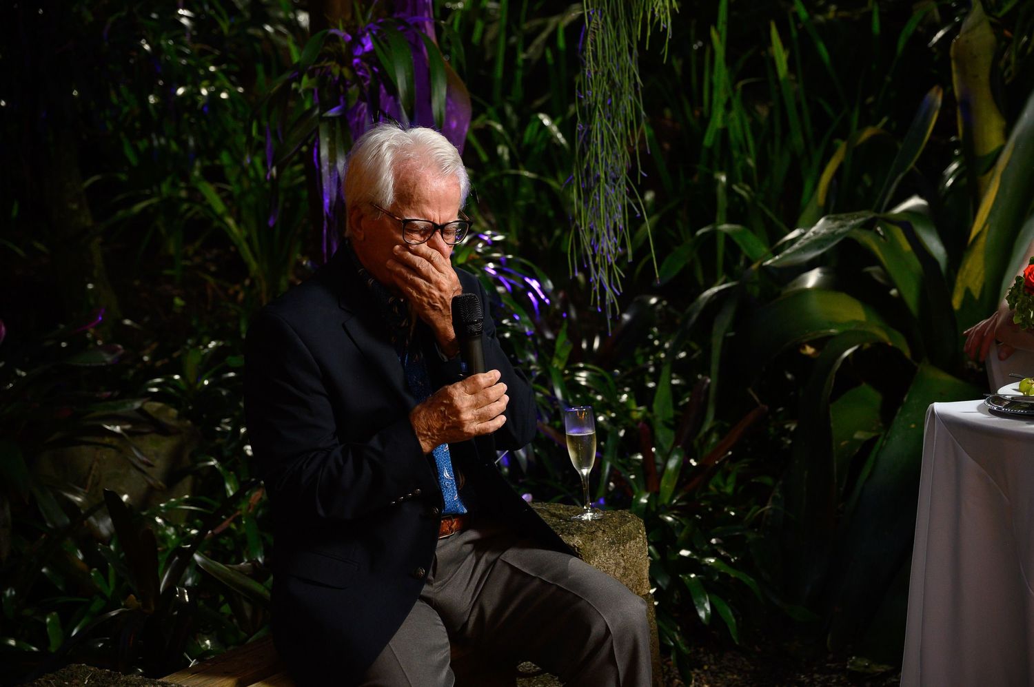 Elderly man in dark blazer sits emotional, covering face with hand, holding champagne glass outdoors at night event.