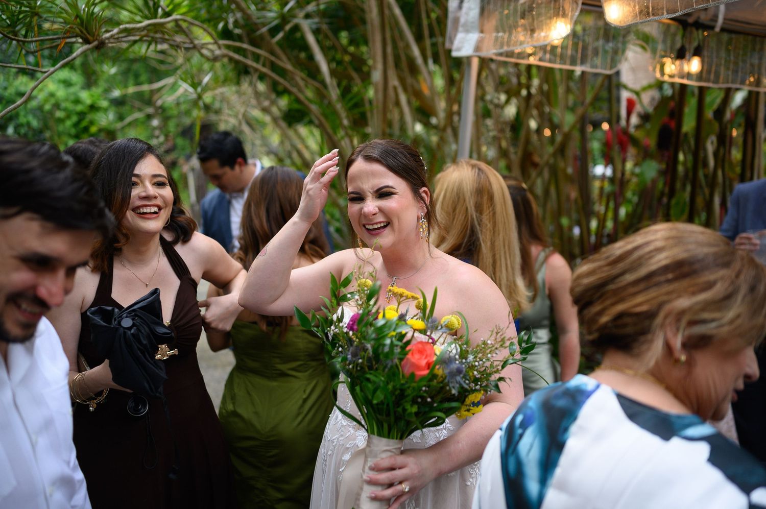Bride holding colorful bouquet laughing with guests at outdoor wedding reception surrounded by lush greenery.