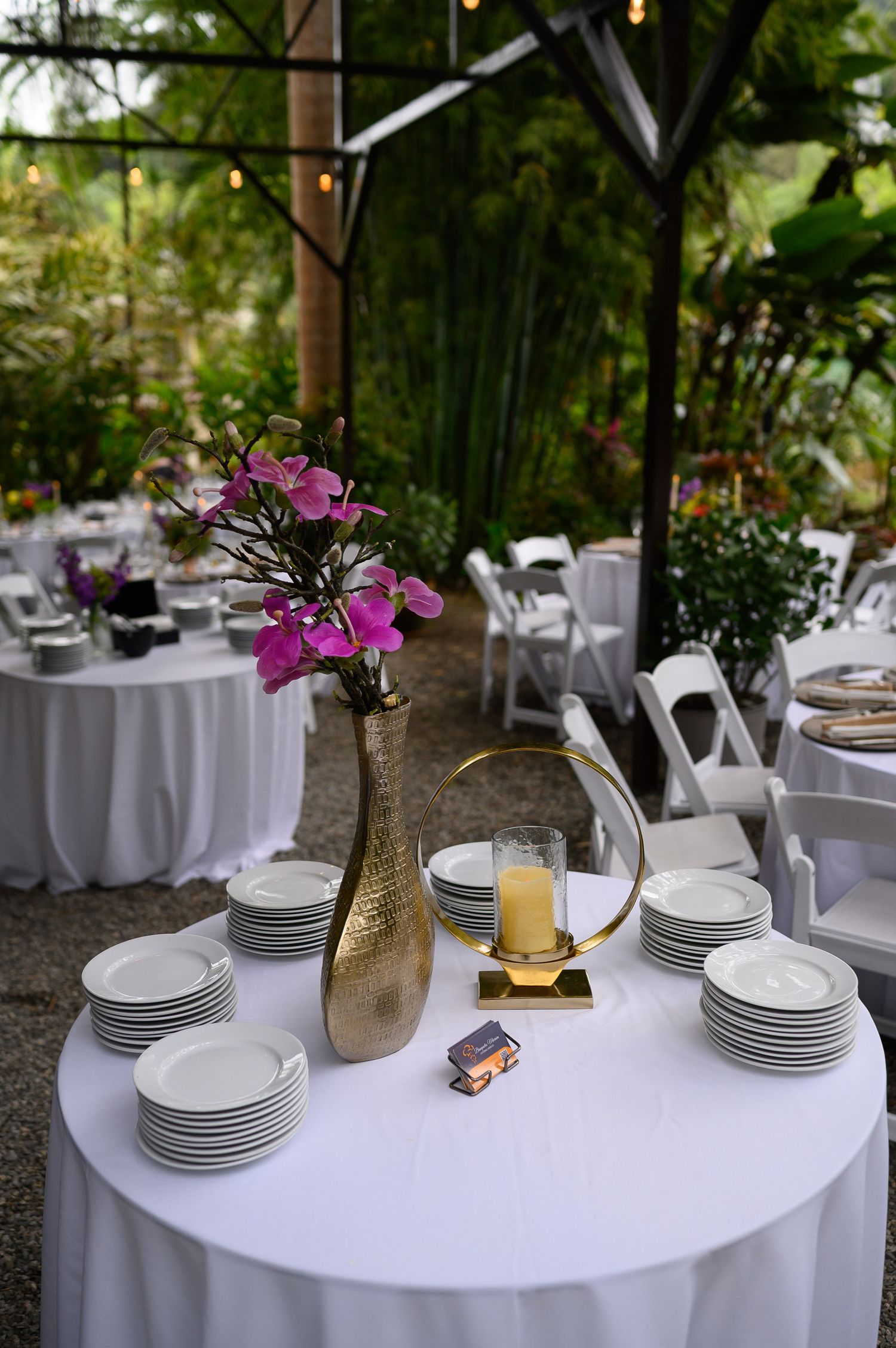 Elegant outdoor wedding reception with white tablecloths, stacked plates, gold vase with pink flowers, and lush greenery backdrop.