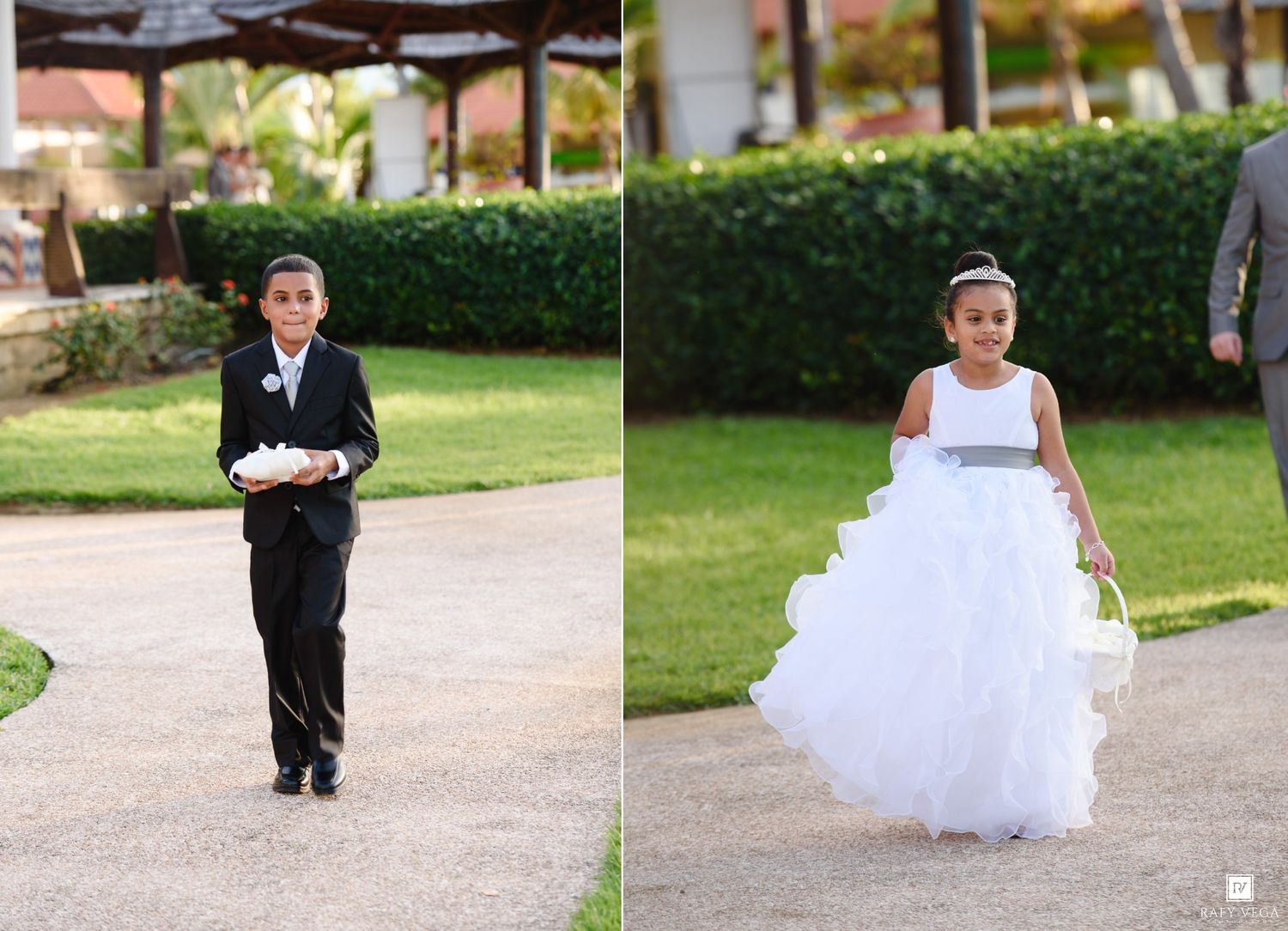 A young ring bearer in a black tuxedo and flower girl in a white dress walk down a path at an outdoor wedding.