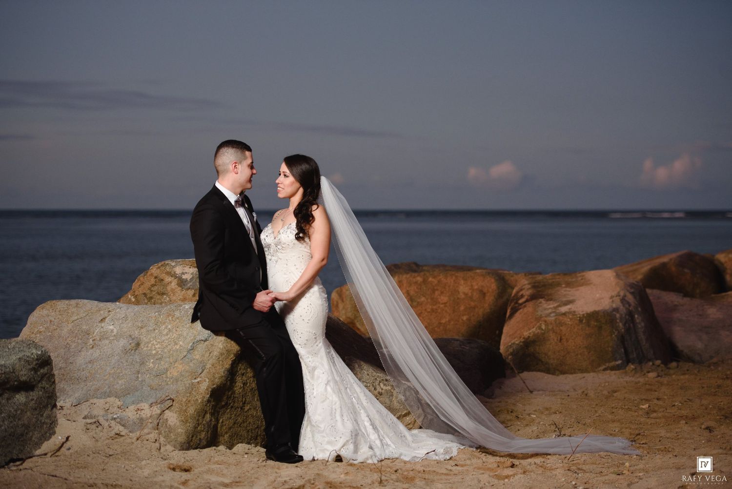 Bride and groom hold hands on rocky beach at sunset, her white mermaid gown and veil flowing in the breeze.