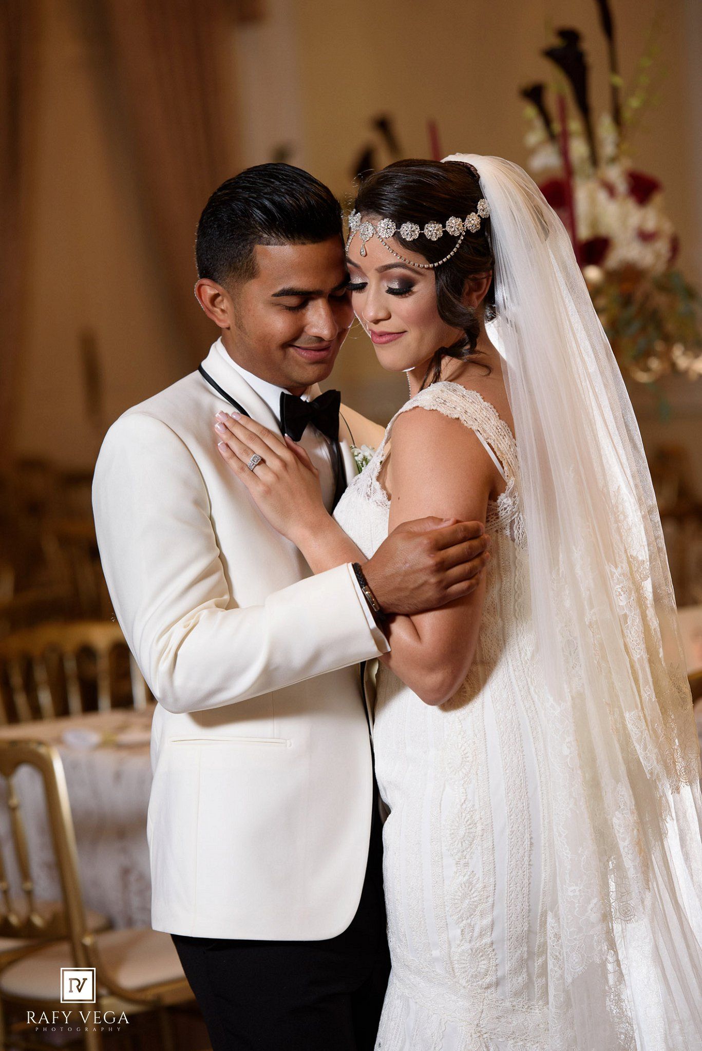 Newlyweds share an intimate moment during first dance, bride in beaded headpiece and lace gown, groom in white jacket.