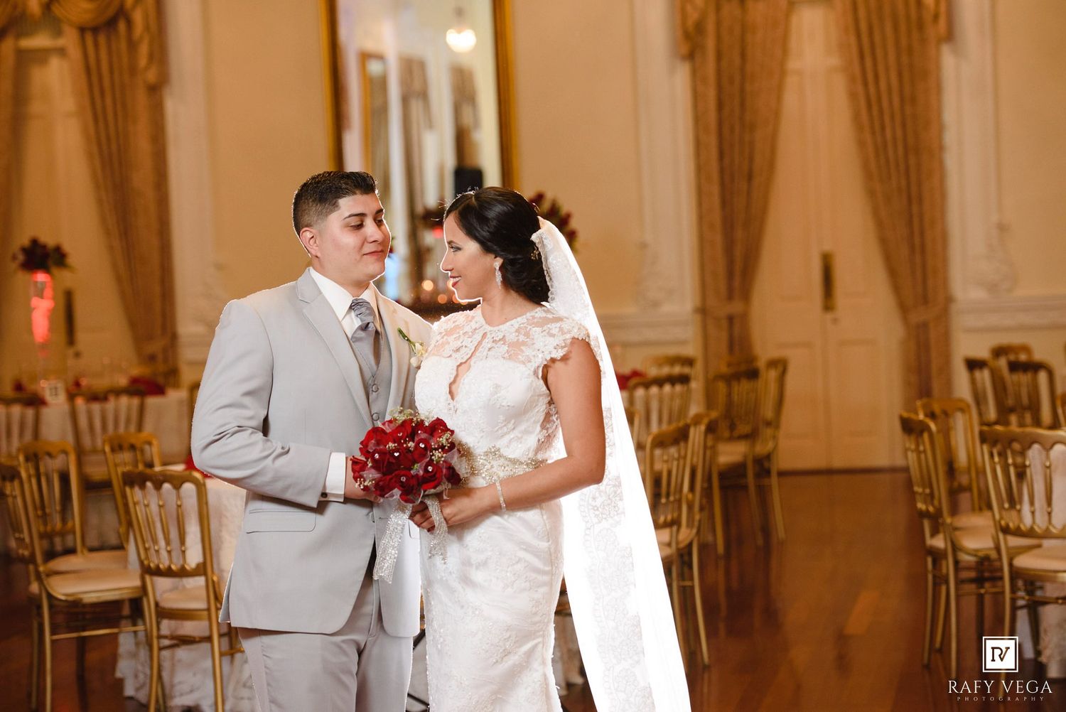 Bride holding red bouquet and groom in gray suit share a tender moment in a gold-chaired wedding venue.