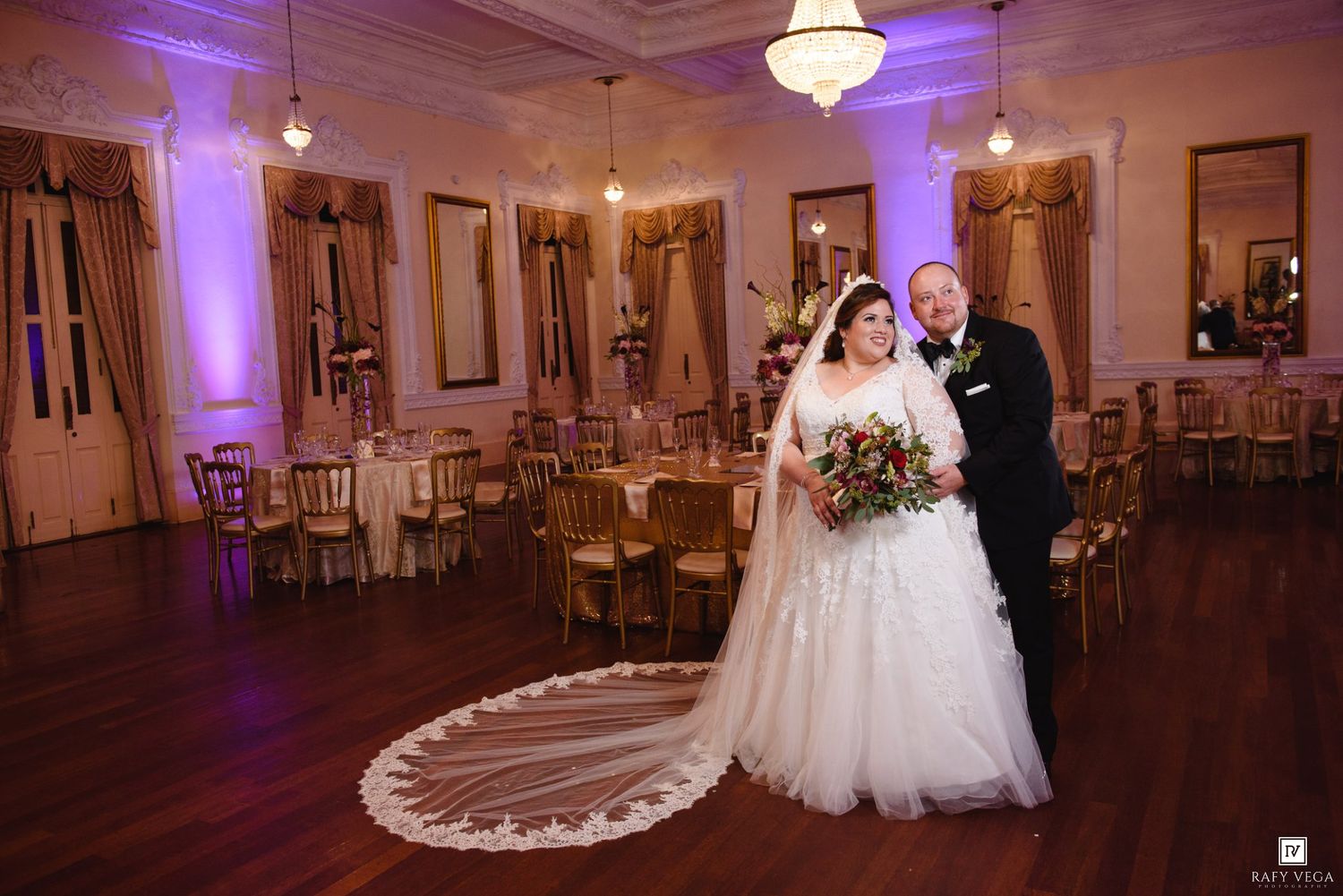 Wedding couple stands in ornate reception hall featuring chandeliers, purple uplighting, and gold banquet seating.