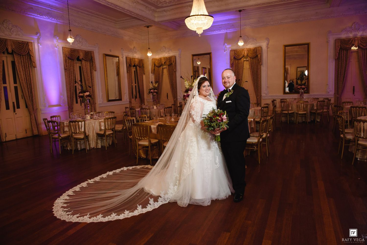 Bride and groom posing in an elegant ballroom with purple uplighting, gold chairs, and a long cathedral veil.