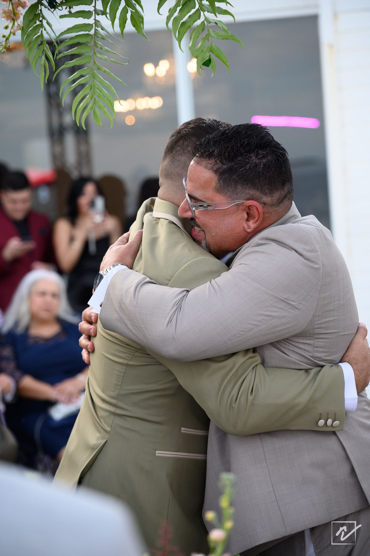 Two men in suits share a heartfelt hug at an outdoor wedding reception while guests watch and take photos in the background.