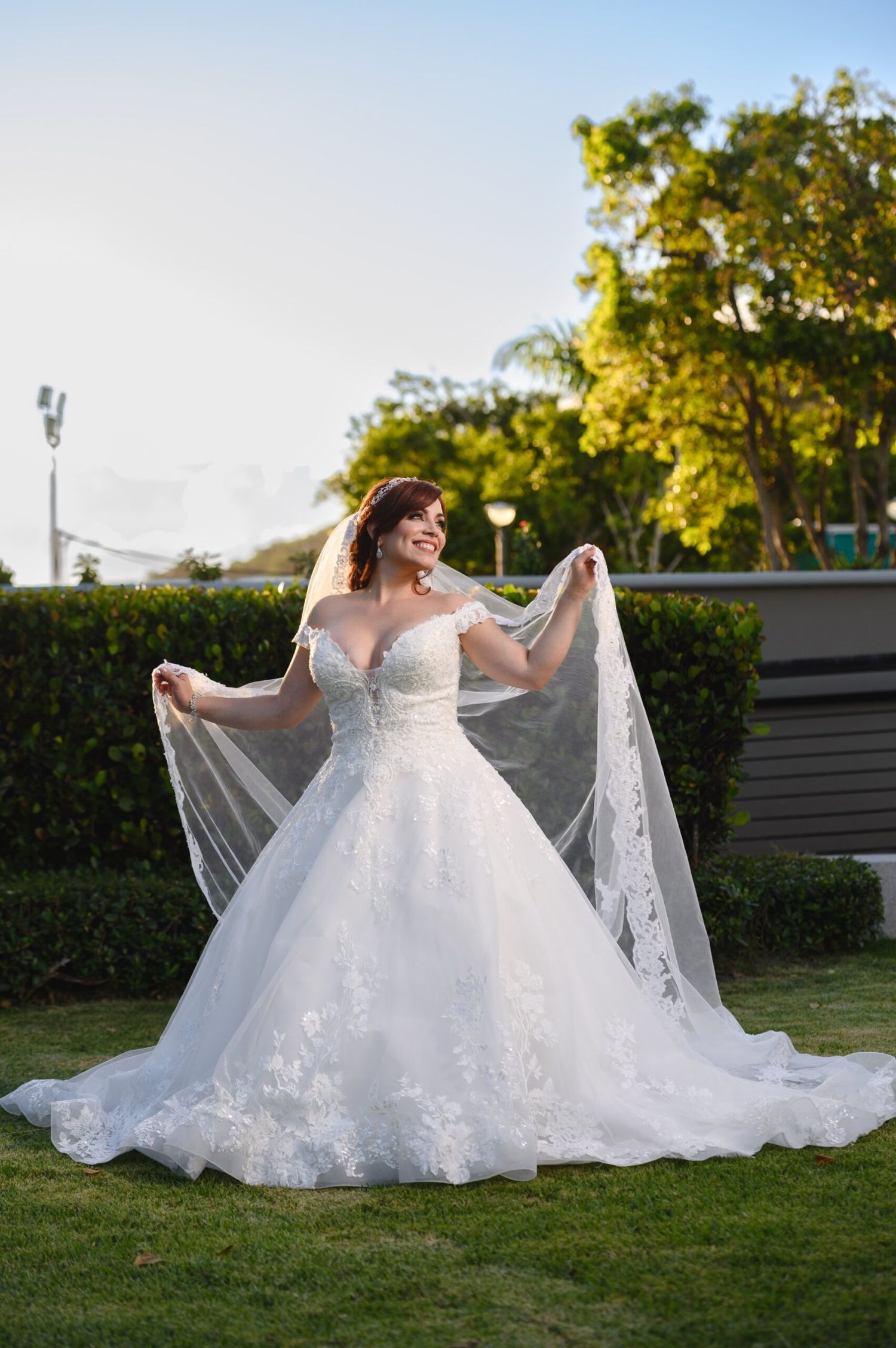 Bride in off-shoulder lace ball gown with long veil spreading wide, smiling outdoors on green lawn.