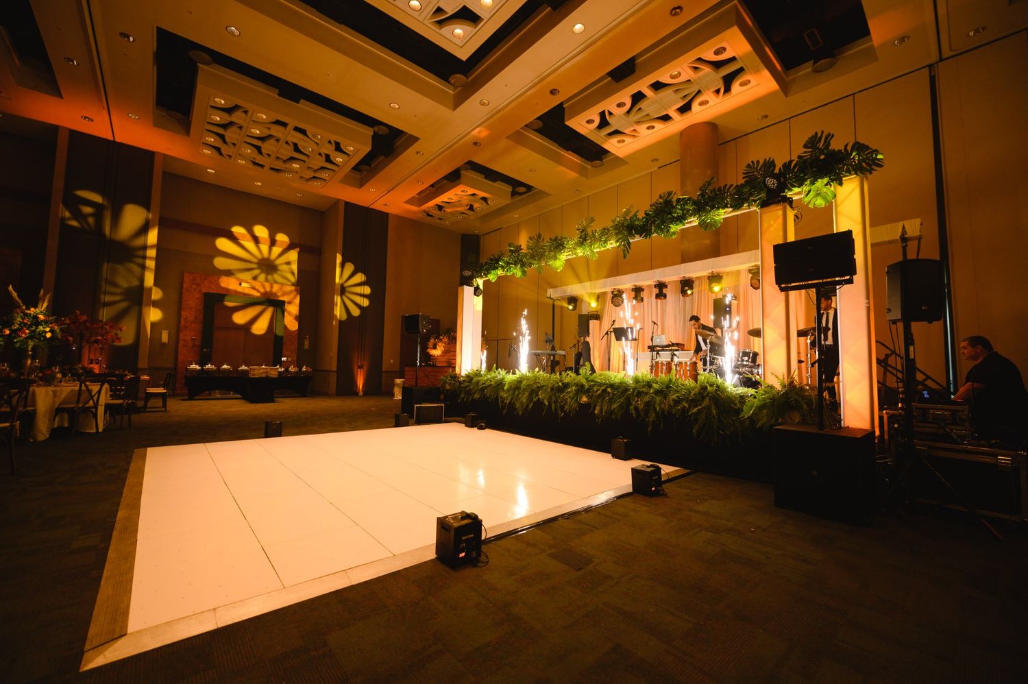 Elegant ballroom with white dance floor, stage with greenery, warm lighting, and decorative ceiling panels.