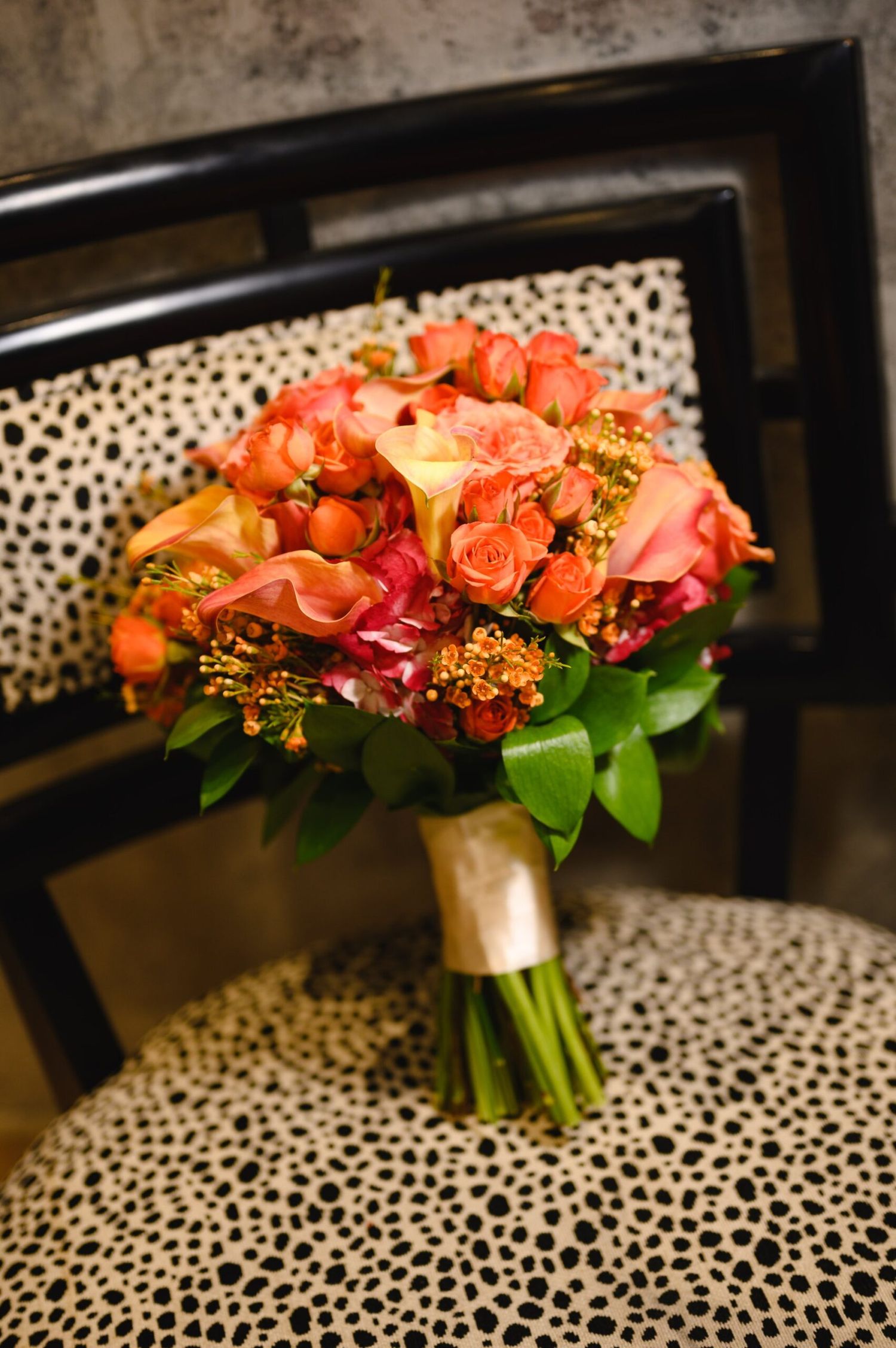 Vibrant bridal bouquet of orange and pink roses with gold accents resting on a black and white spotted chair.