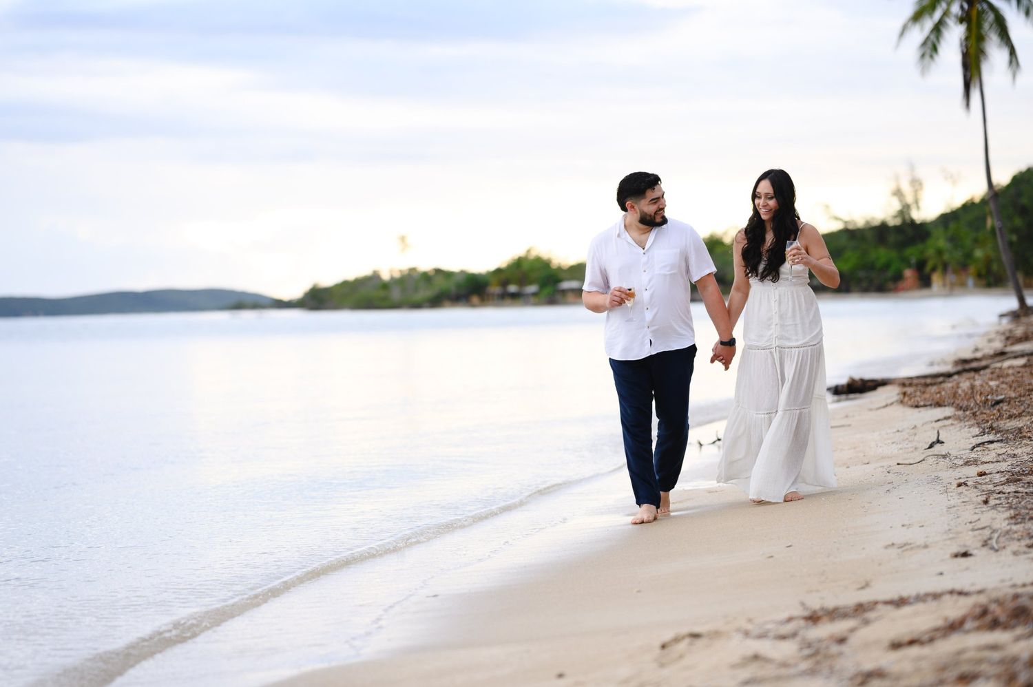 Couple holding hands walking on a tropical beach at sunset, man in white shirt and navy pants, woman in white dress.
