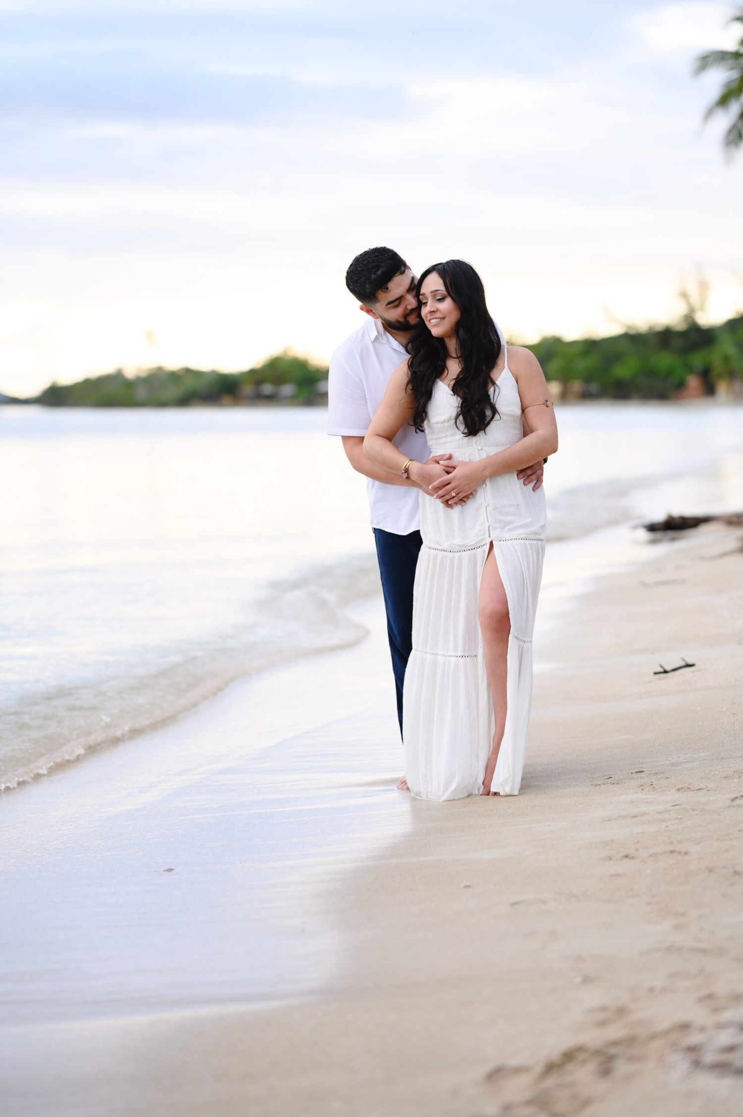 Couple embracing on a sandy beach at sunset, woman in white maxi dress and man in white shirt and navy pants.
