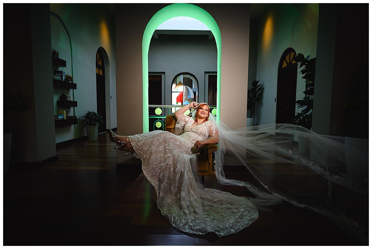 Bride in lace wedding gown and veil poses elegantly on floor in dramatic lit venue with arched doorways.