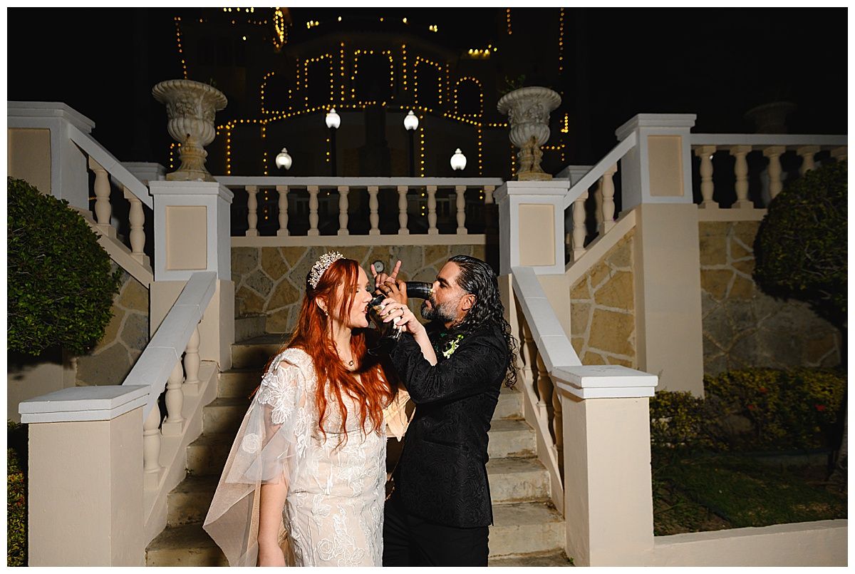 Bride and groom share a romantic toast outdoors at night, surrounded by elegant stone stairs and warm fairy lights.
