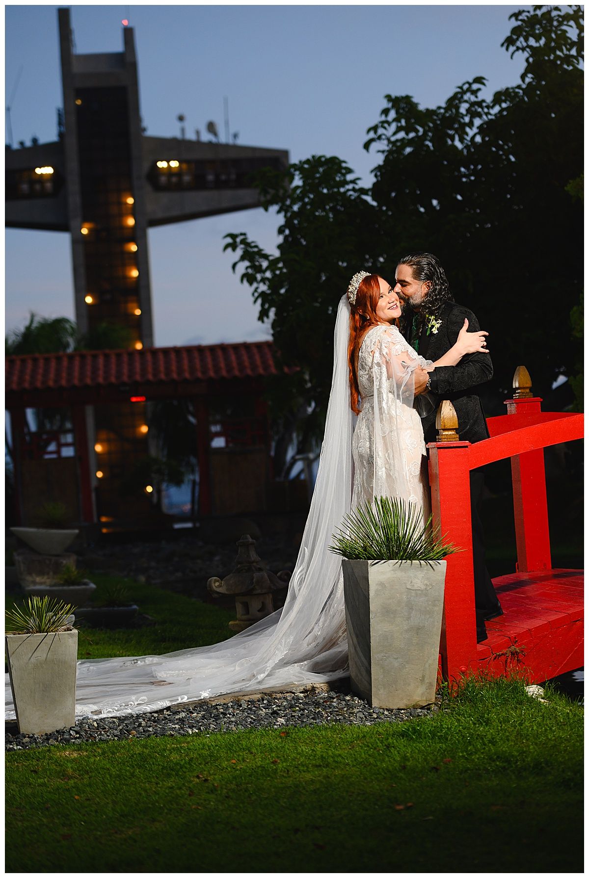 Bride and groom share a romantic moment on a red bridge at dusk, with a long white veil and garden lights behind them.
