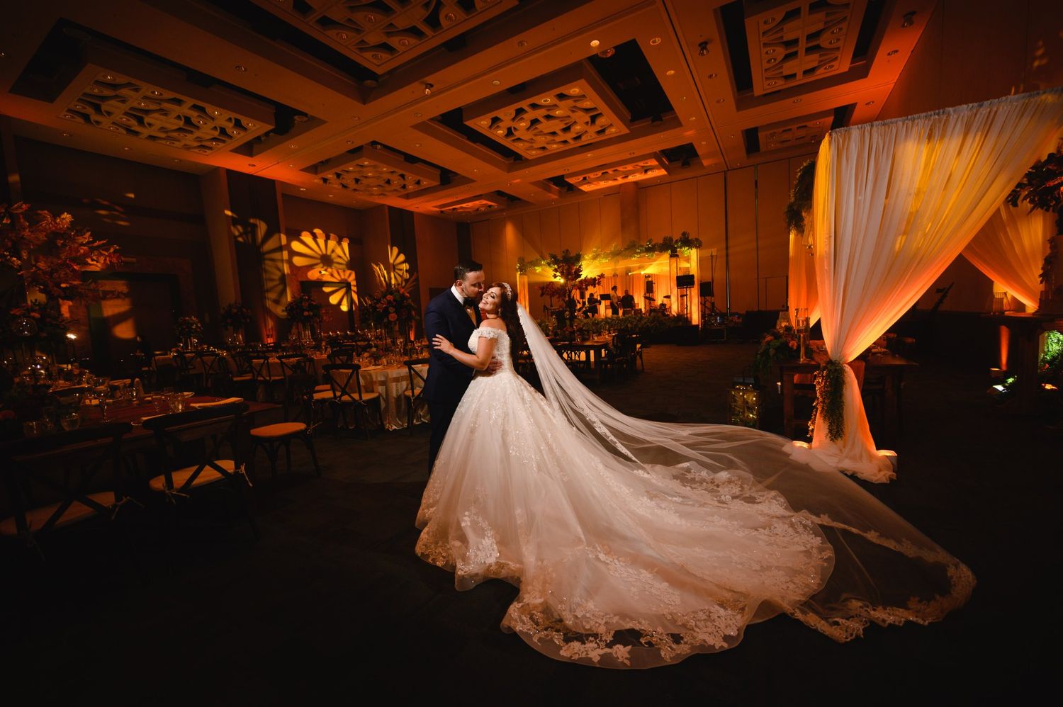 Bride and groom share first dance in elegant ballroom with warm amber uplighting and flowing white wedding gown train.