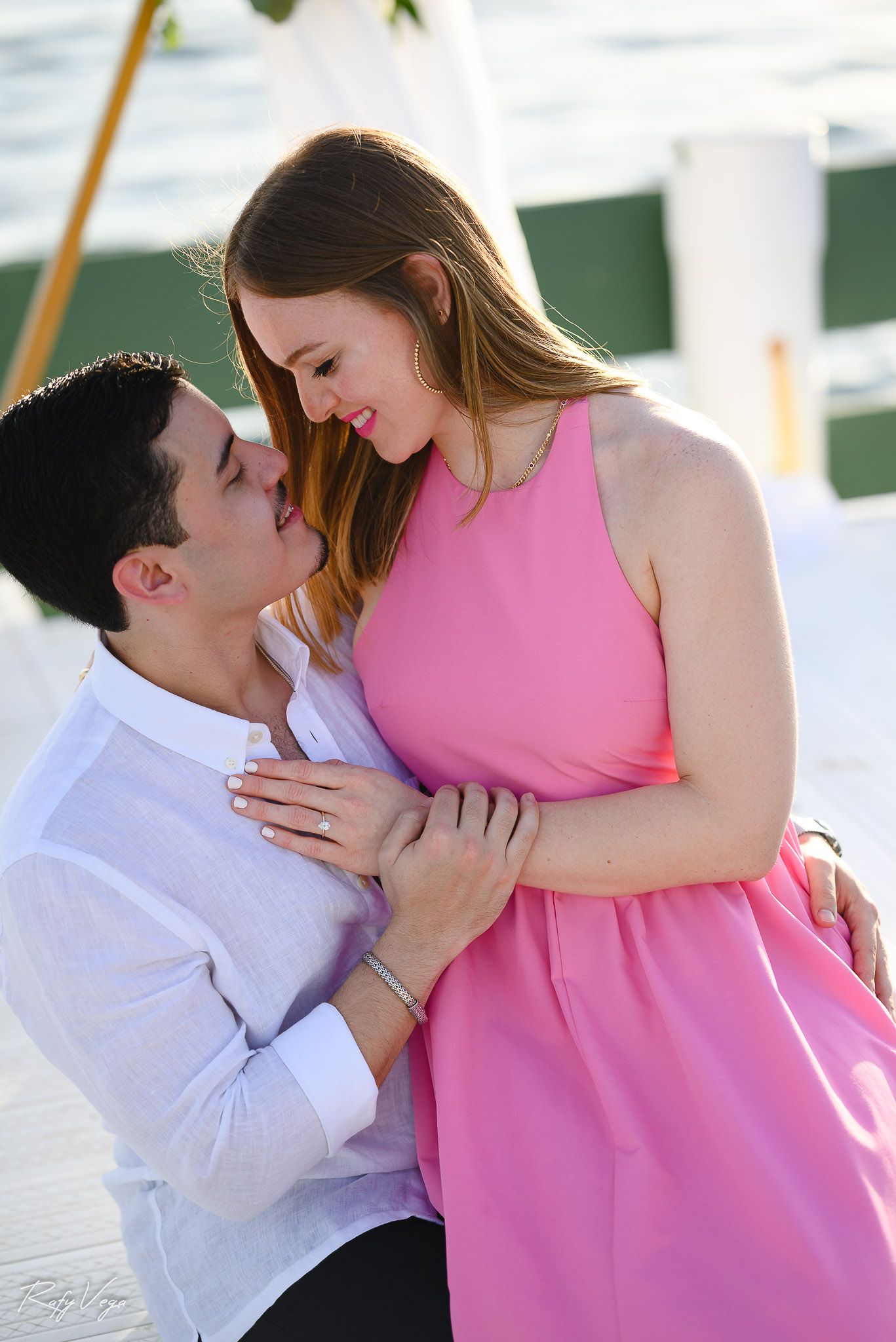 Couple sharing a romantic moment on a waterfront dock, woman in pink dress and man in white shirt embracing.
