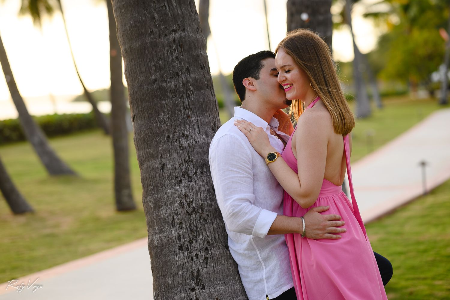 Couple embracing romantically near a palm tree in a tropical park at sunset, woman in pink dress.
