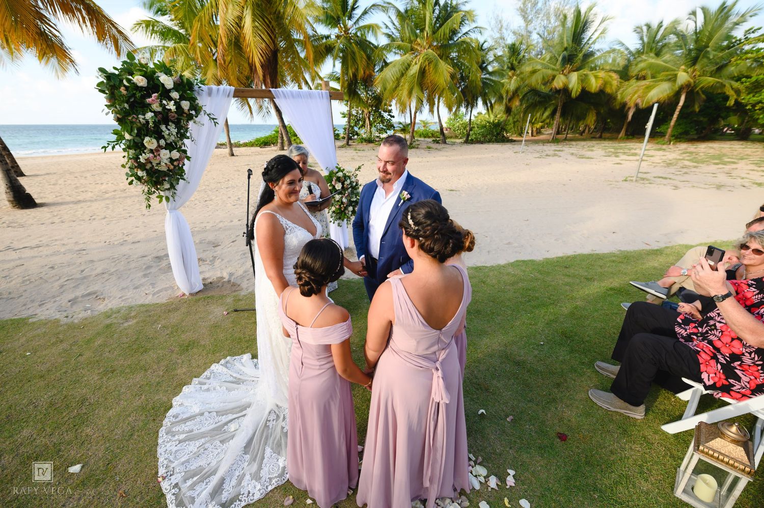 Bride and groom exchange vows at a tropical beach wedding ceremony with bridesmaids in pink dresses and palm trees.