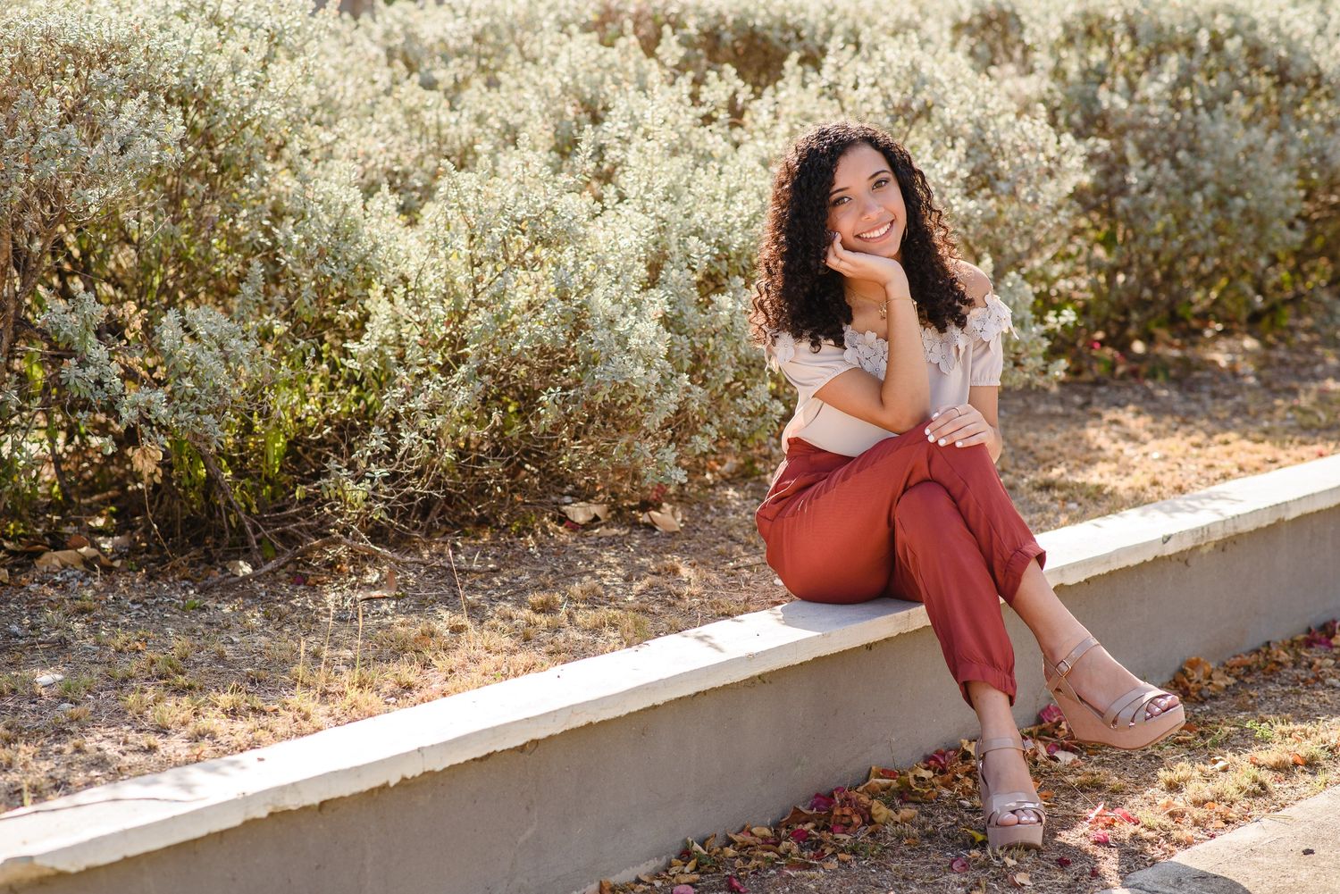 Young woman with curly hair smiling, wearing rust pants, seated on a concrete ledge outdoors.