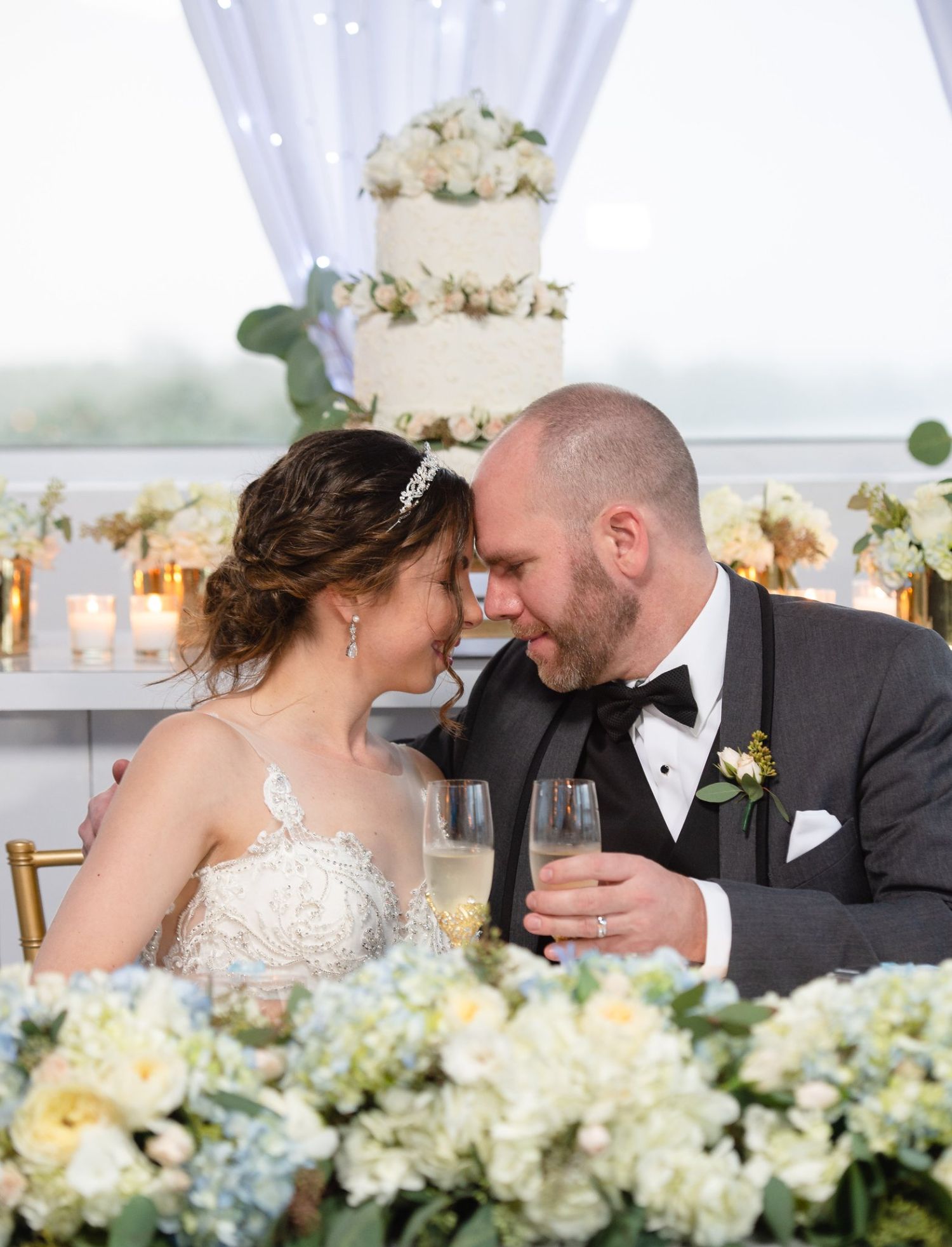 Bride and groom sharing an intimate moment at sweetheart table with wedding cake and floral arrangements behind them.