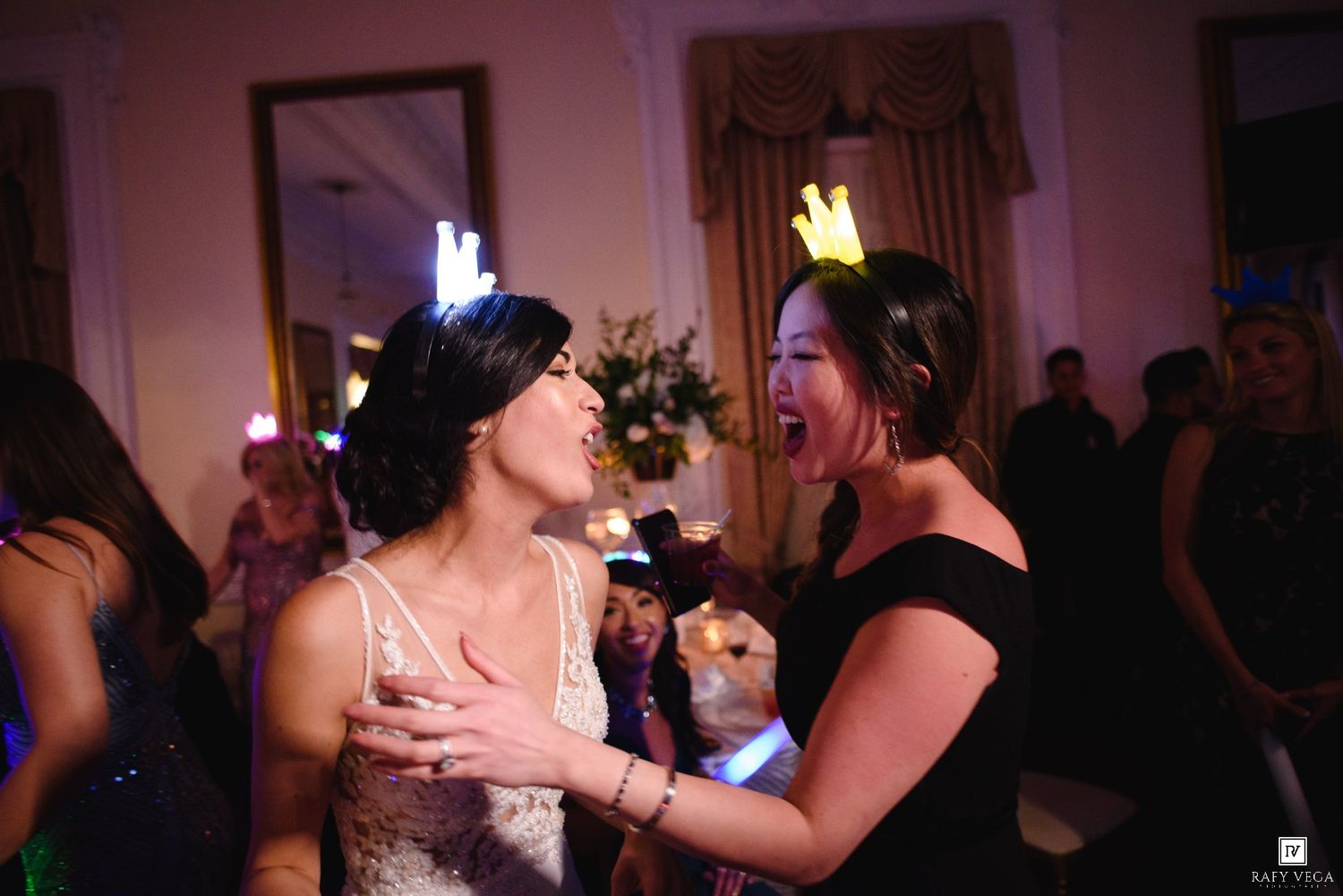Two women laugh and dance together at a wedding reception, one in a white bridal gown and the other in a black dress.