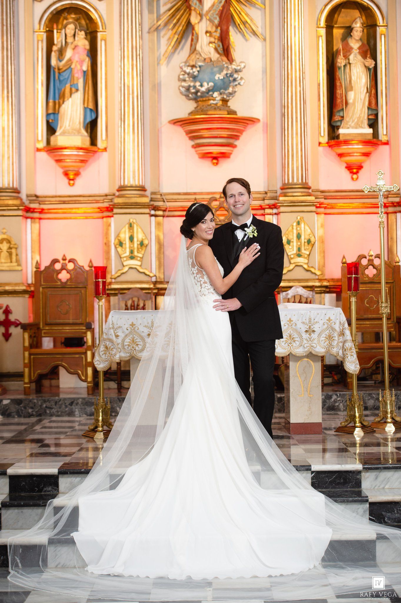 Bride in elegant gown with long veil poses with groom before golden church altar with religious statues.