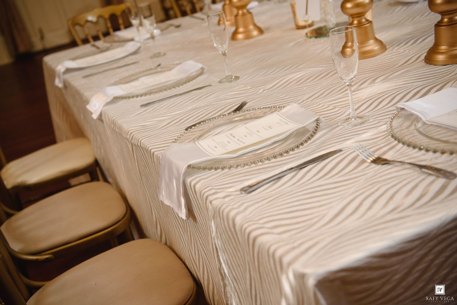 Elegant wedding table setting with ivory wave-patterned tablecloth, white napkins, silver cutlery, and gold candle holders.