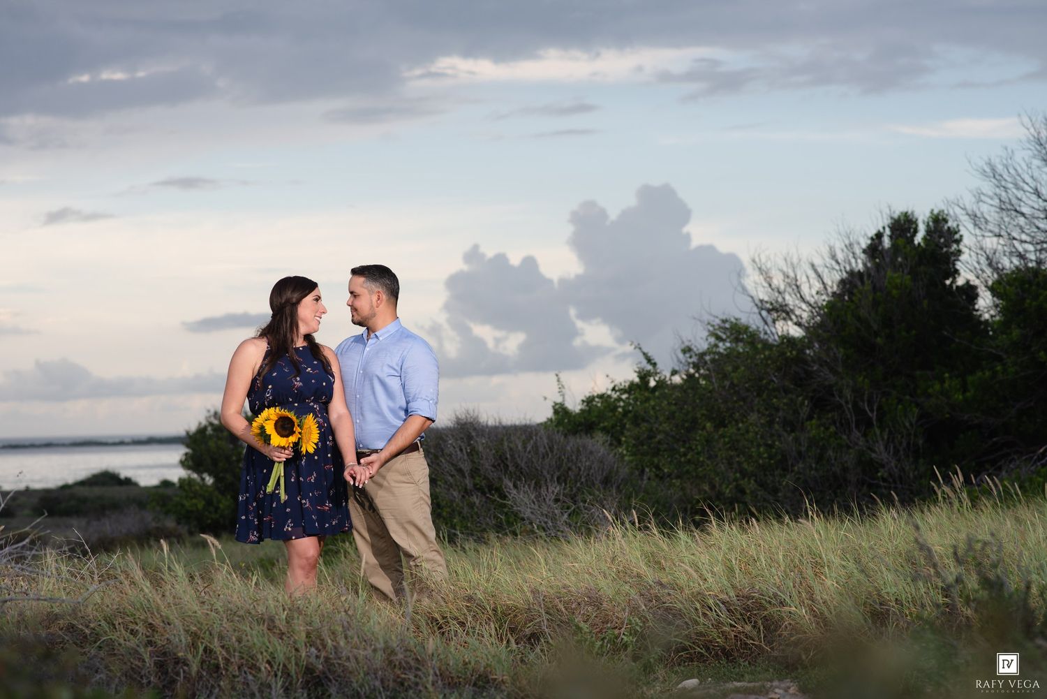 Couple holds hands in a grassy field at sunset, woman in navy dress with sunflower bouquet, man in light blue shirt.