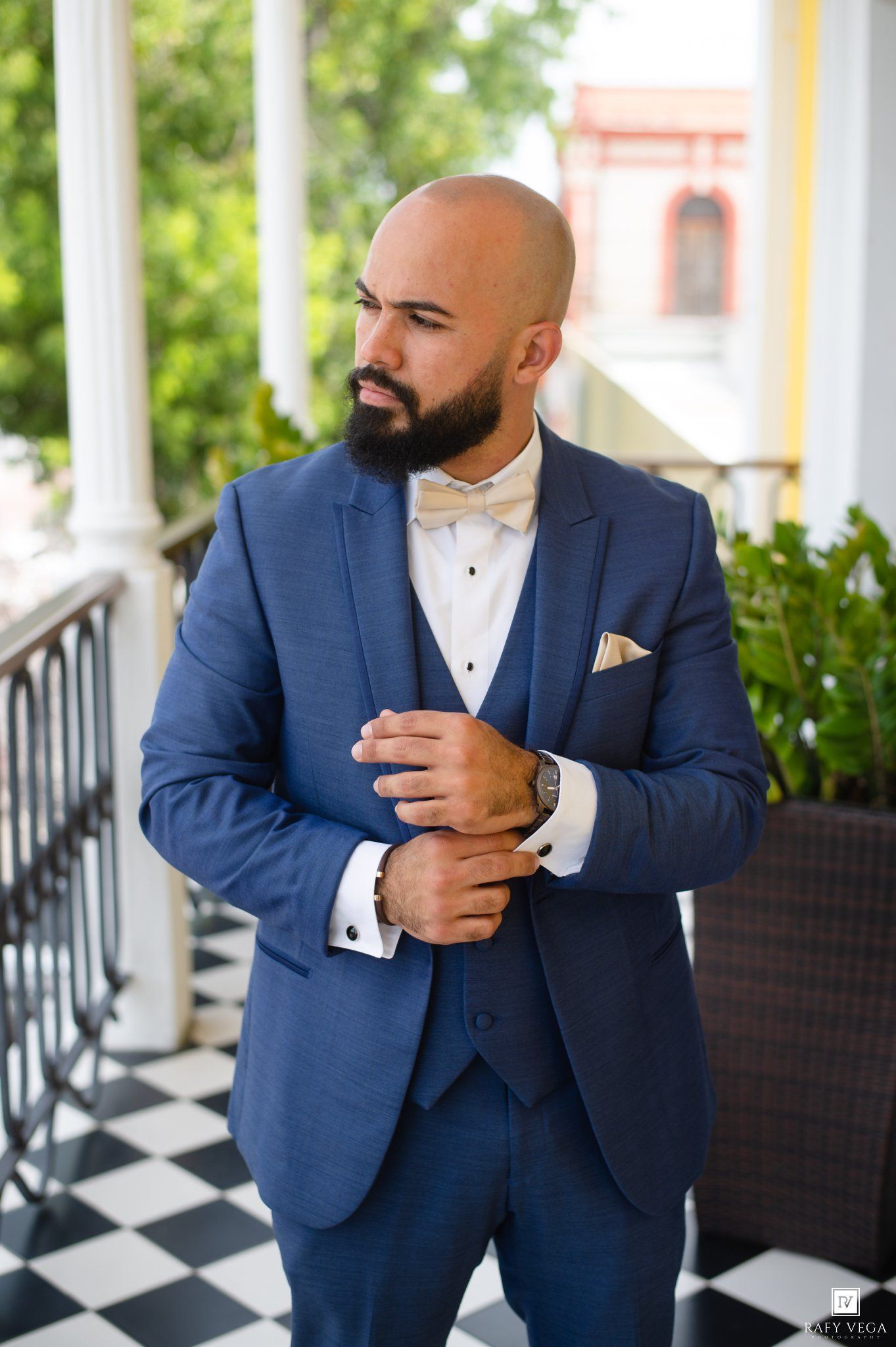 Bald groom with beard adjusting cufflinks in a navy blue three-piece suit with gold bow tie on a checkered floor porch.