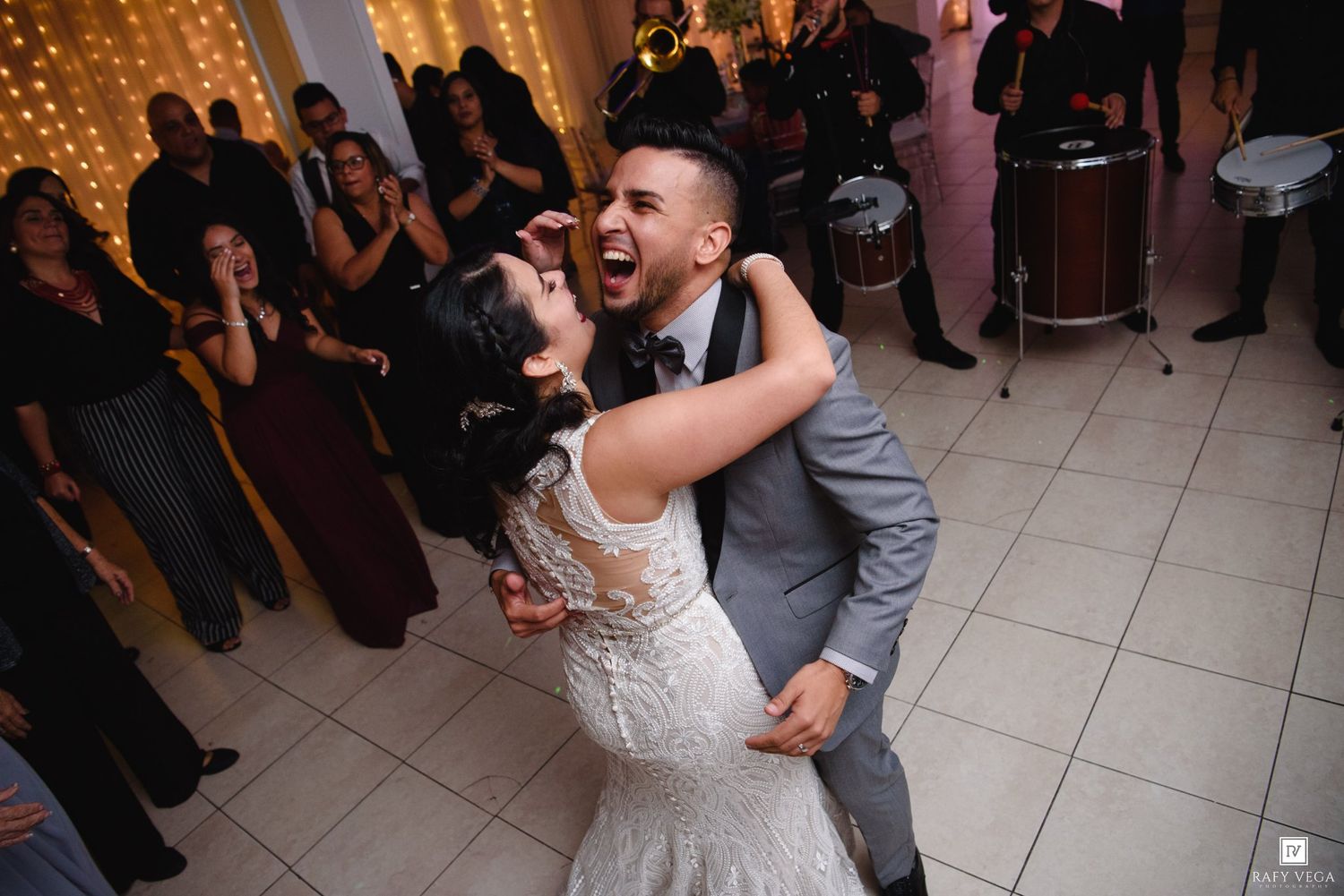 Joyful bride and groom share first dance at wedding reception with guests watching and conga drums in background.