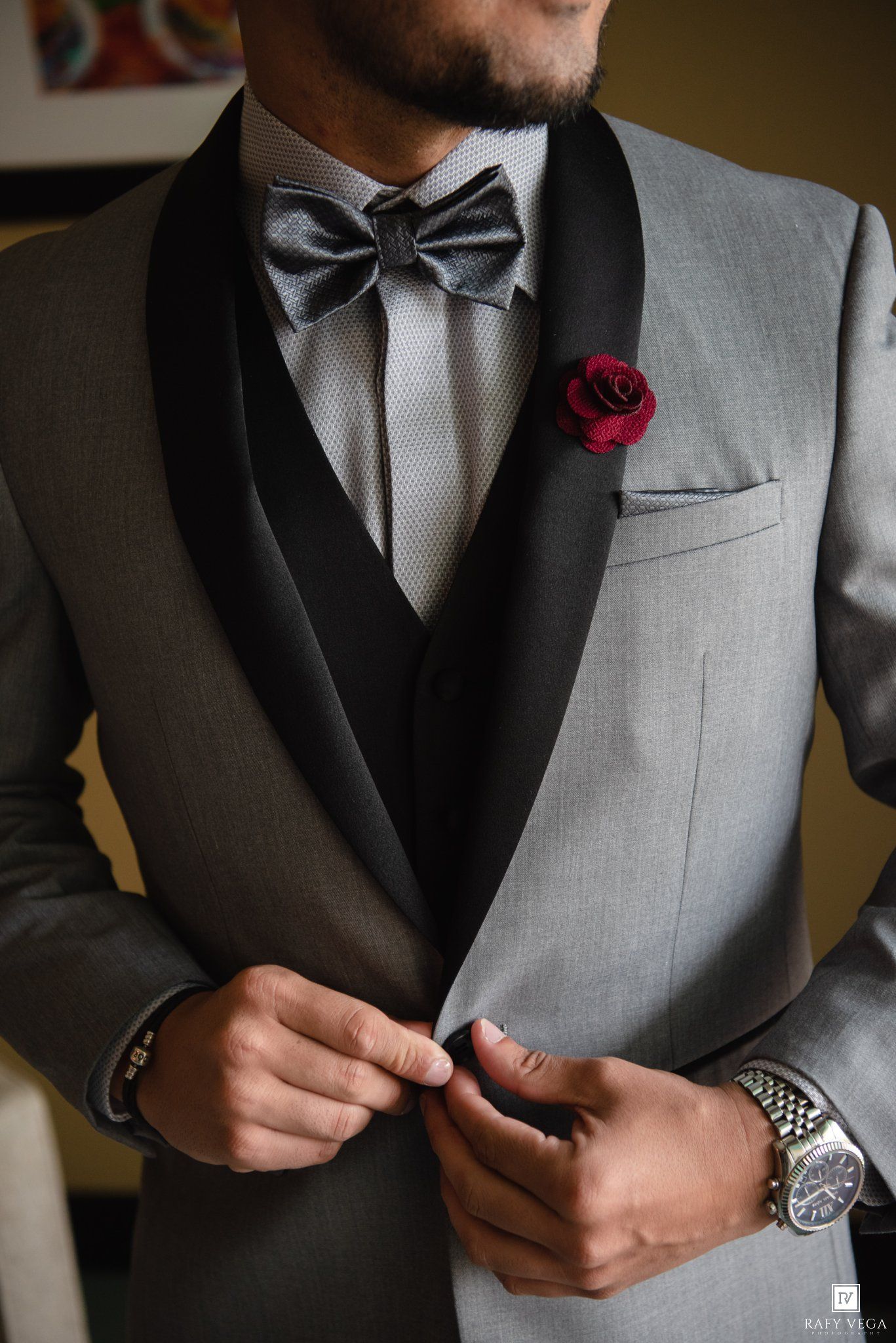 Man in gray tuxedo with black lapels, bow tie, red rose boutonniere, and silver watch buttoning jacket.