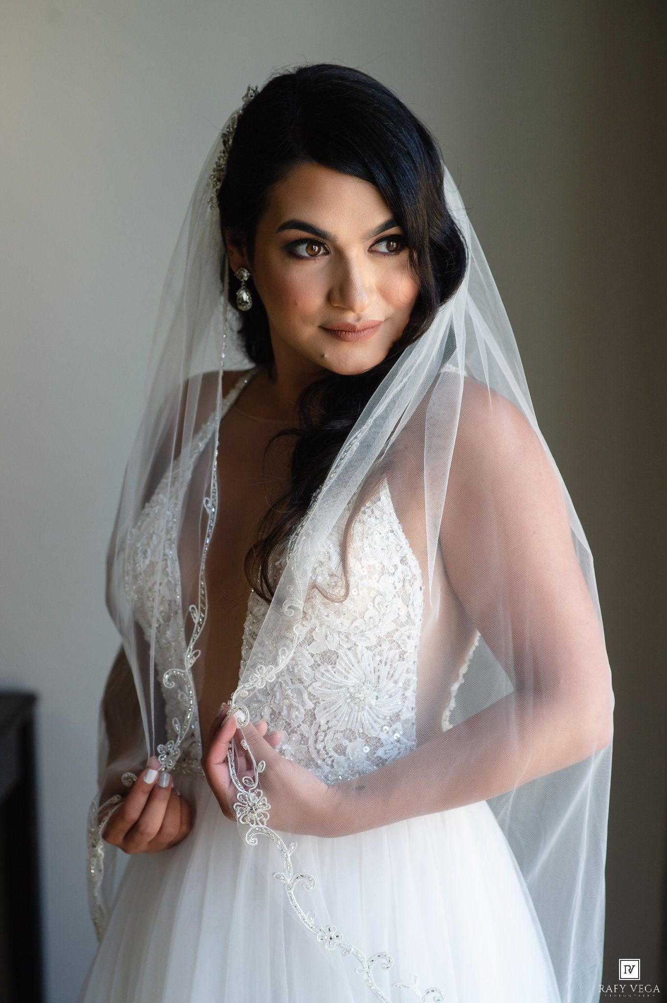 Beautiful dark-haired bride in lace gown and cathedral veil gazes over shoulder in a stunning portrait.
