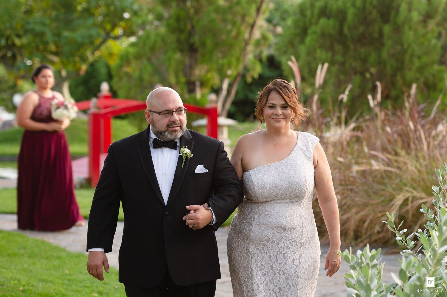Bride in silver lace gown walking arm in arm with groom in black tuxedo at outdoor ceremony.