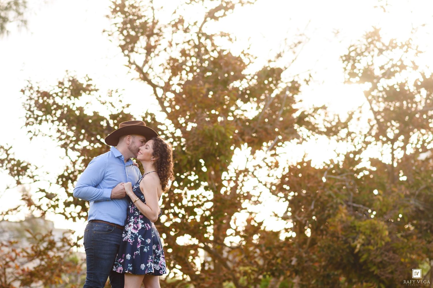 Couple sharing a romantic kiss outdoors beneath a sunlit autumn tree with colorful foliage.