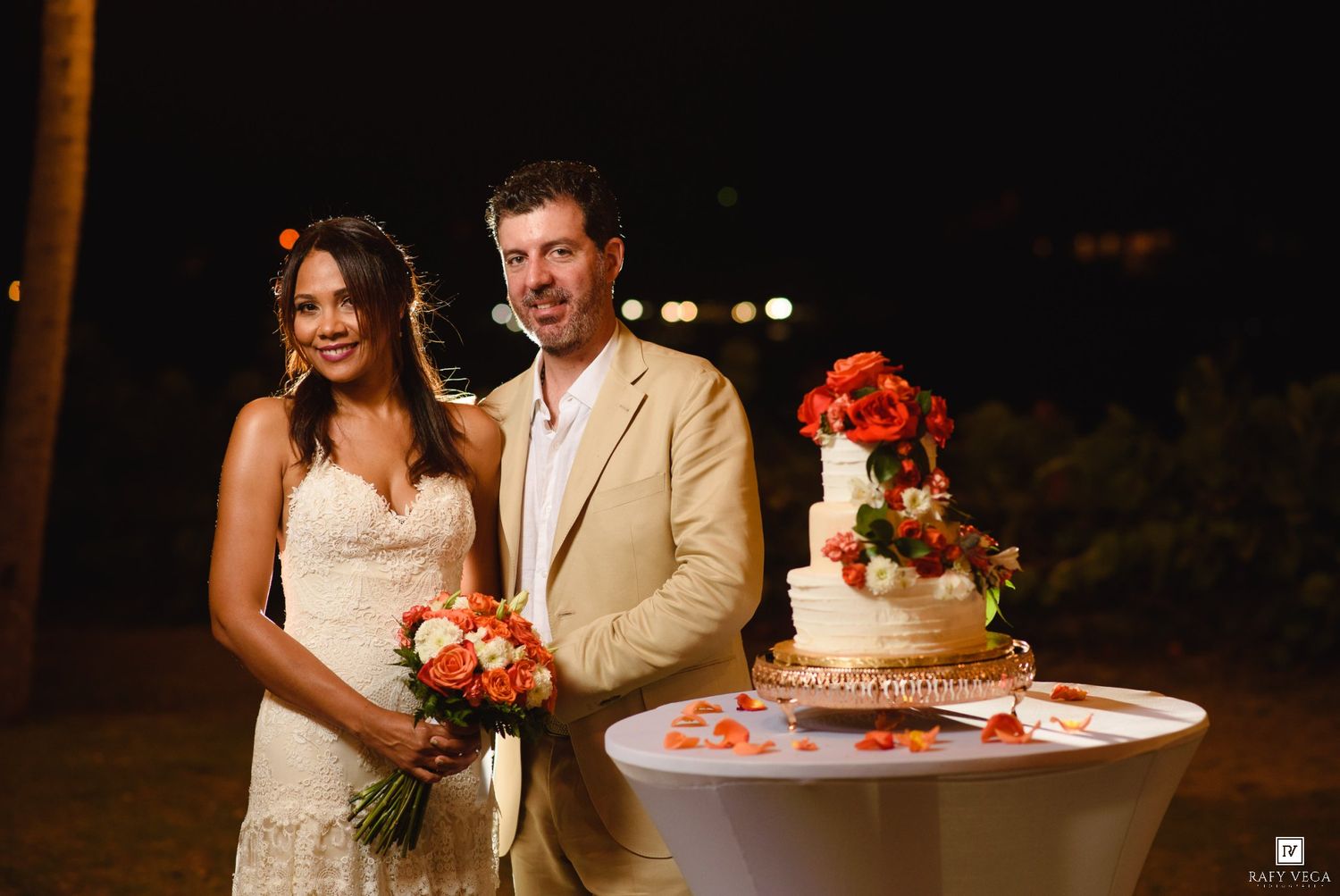 Couple poses beside a tiered wedding cake with orange florals at an outdoor evening reception.