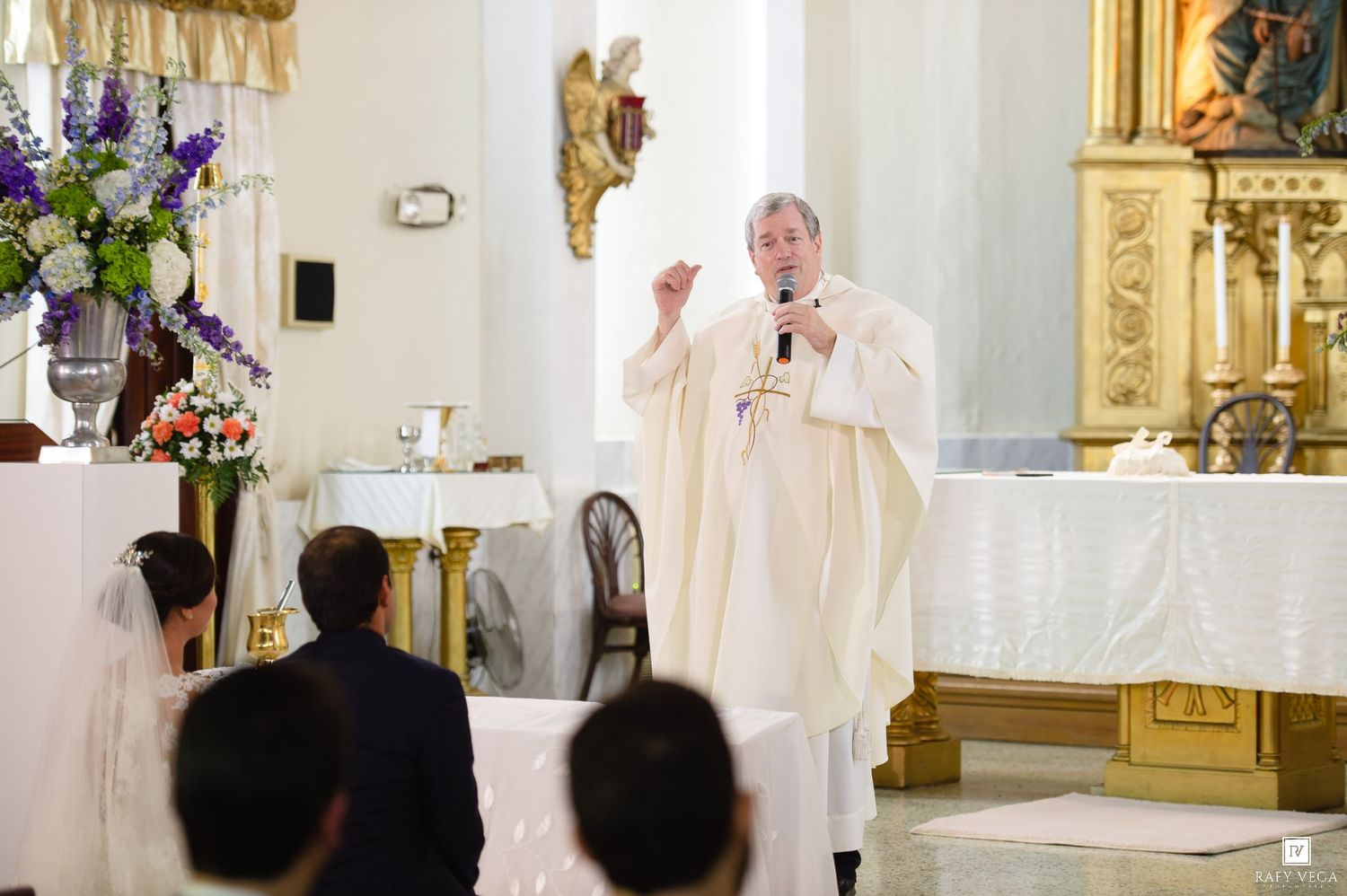 Priest in white vestments speaks at church altar during wedding ceremony with floral arrangements nearby.