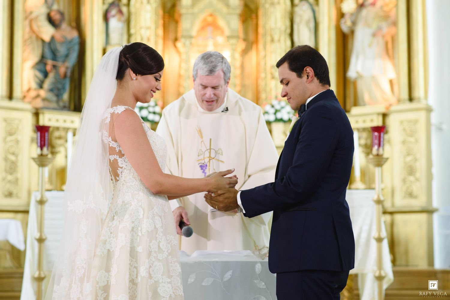 Bride and groom exchange rings at church altar with officiant during elegant wedding ceremony.